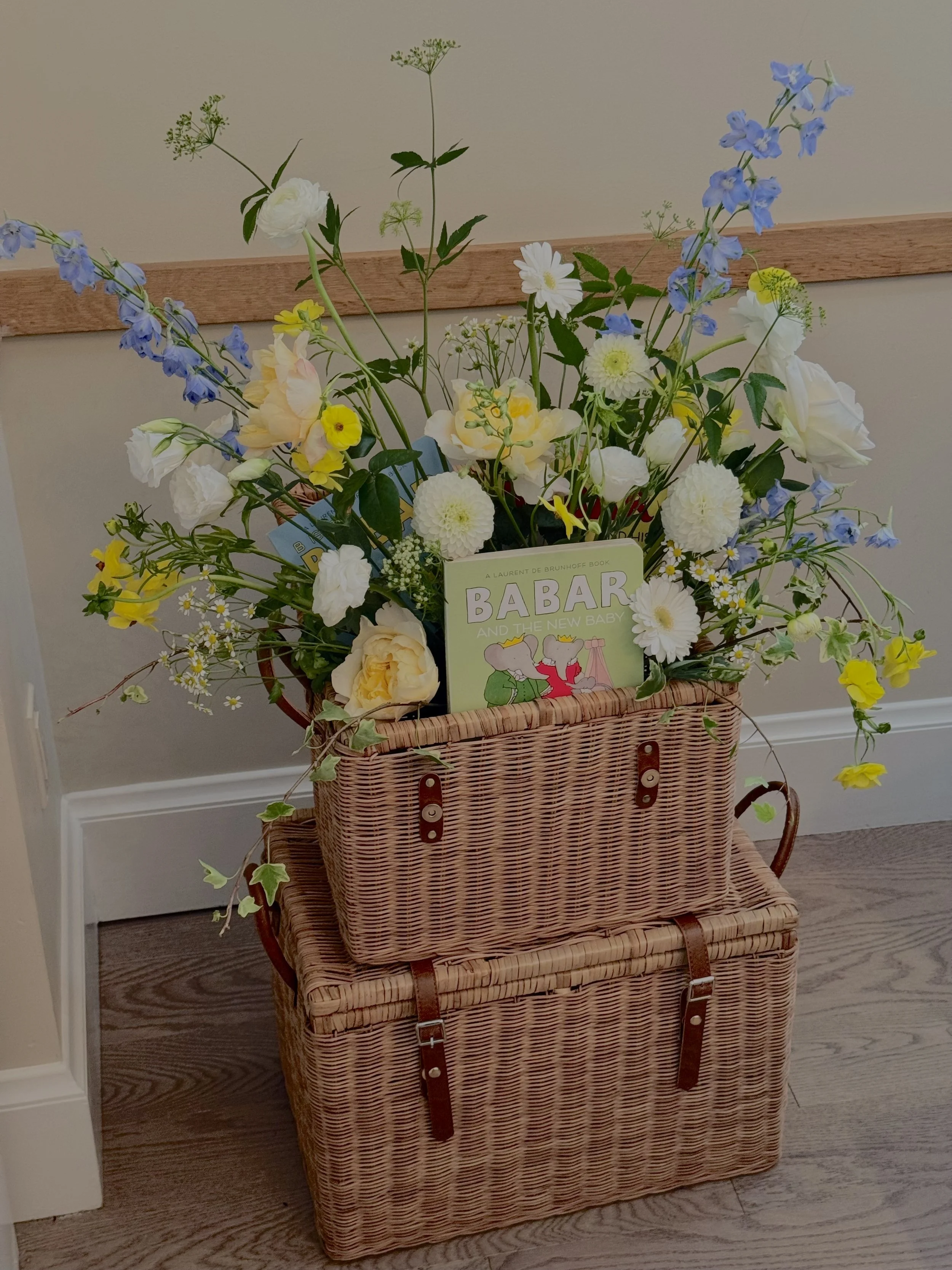 A wicker basket filled with colorful flowers, including white, yellow, and blue blossoms, placed on a wooden table with a small book titled 'BABAR' resting among the flowers.