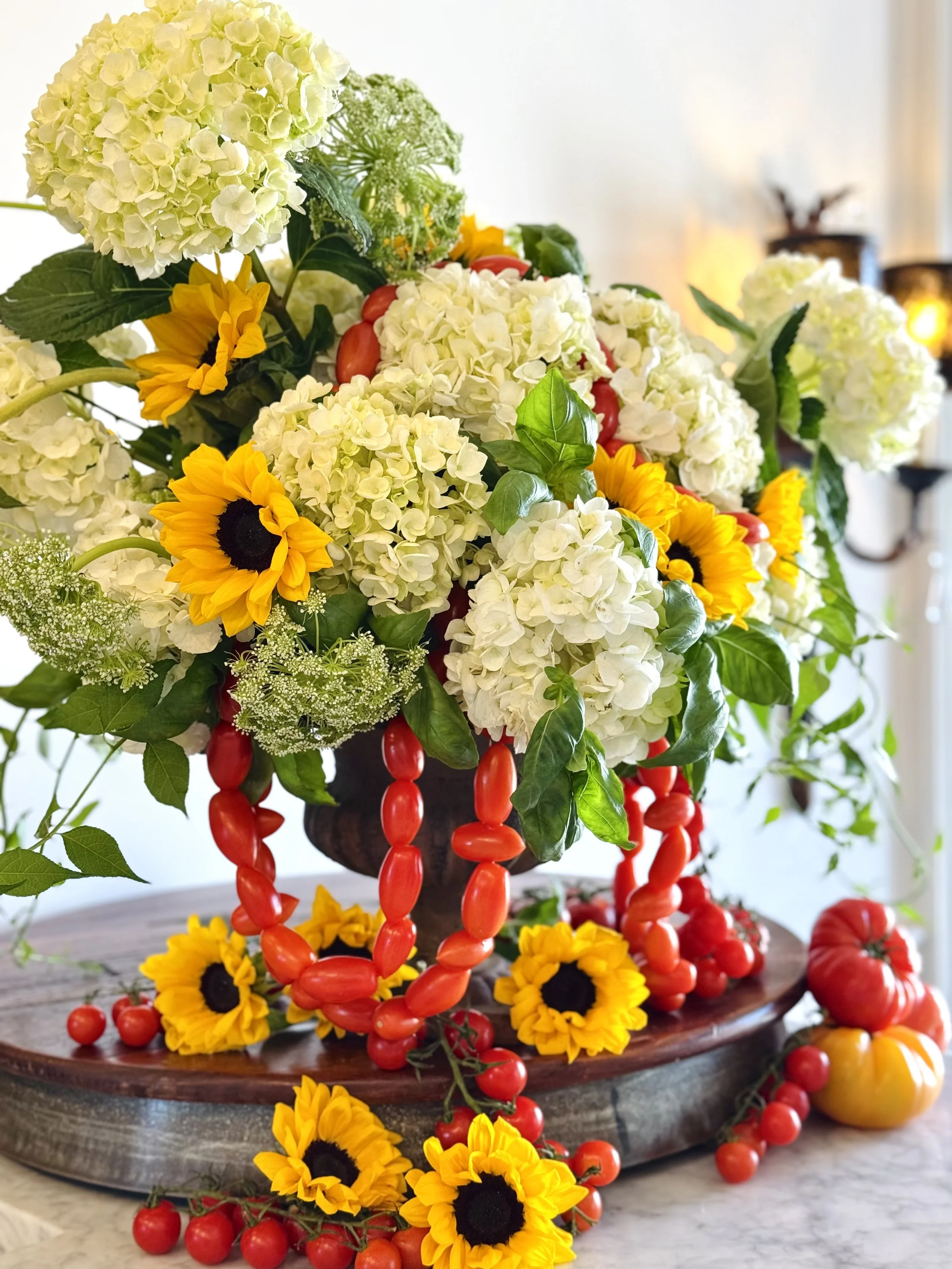 A floral arrangement with white hydrangeas, yellow sunflowers, and green leaves on a wooden table, with red cherry tomatoes and heirloom tomatoes cascading from the arrangement.