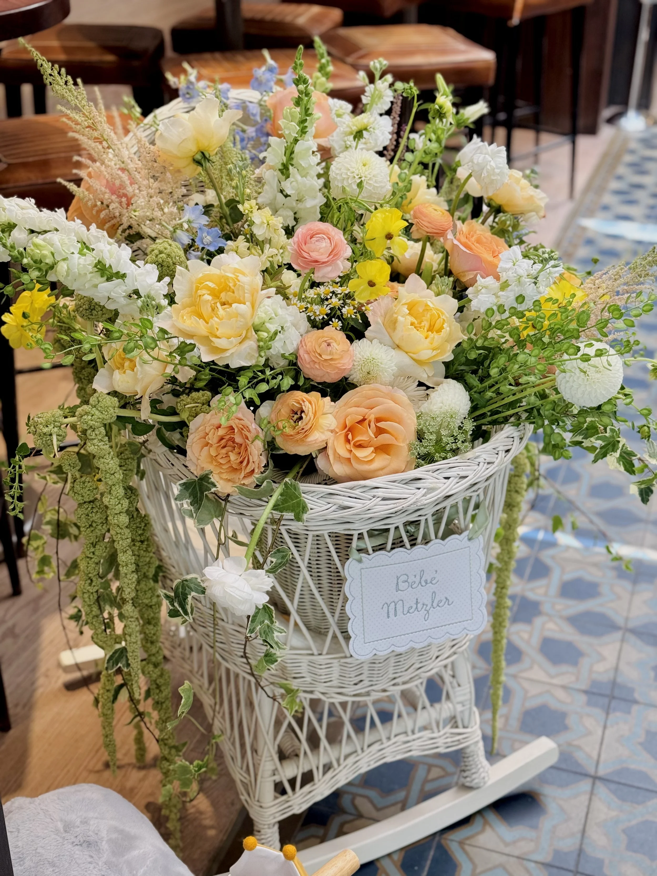 A white wicker basket filled with various pastel-colored flowers including roses, ranunculus, and delphiniums, with greenery and ivy draping over the sides, on a patterned tile floor. There is a handwritten sign on the basket that reads 'Bébé Metzler