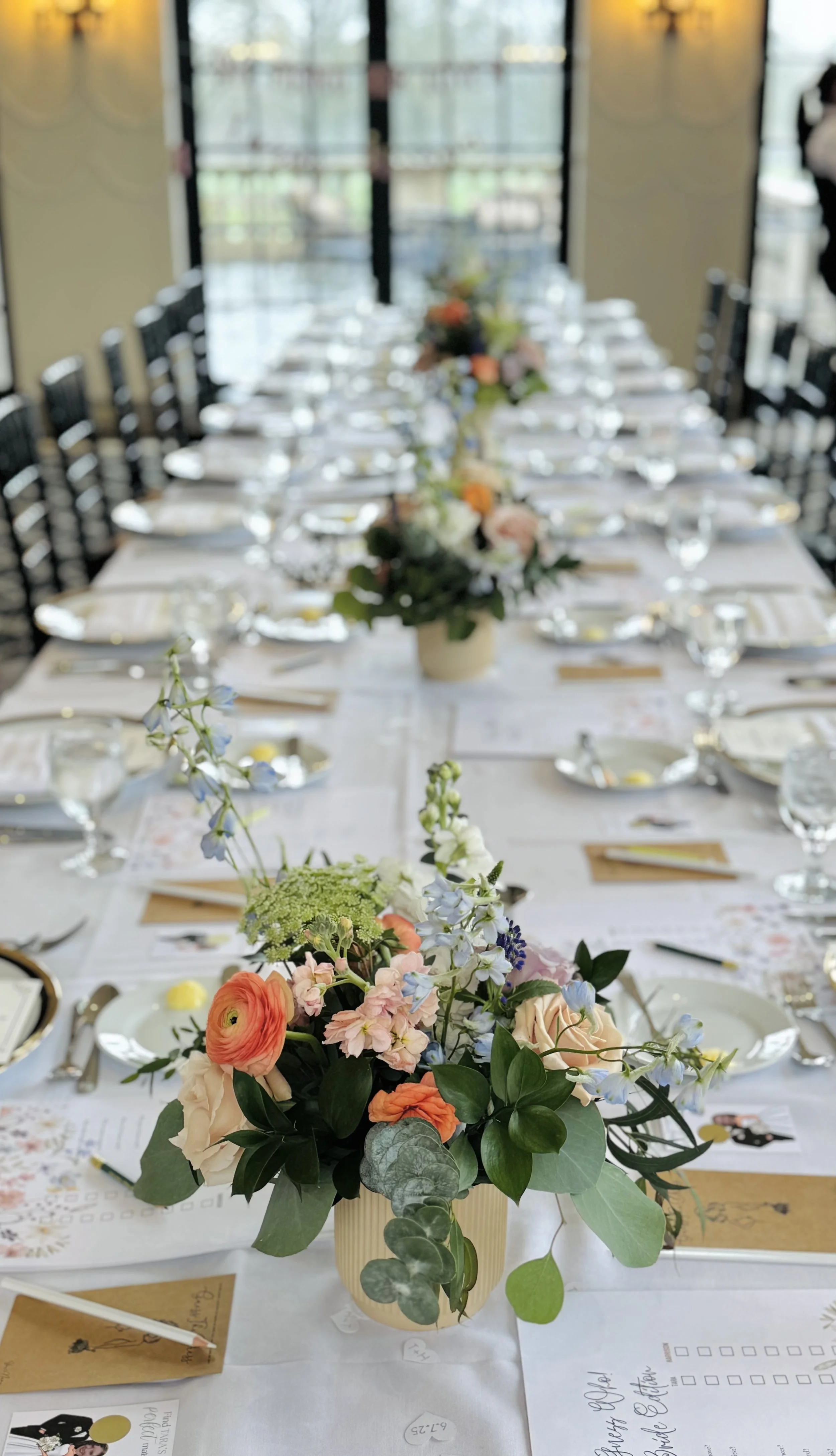 A floral centerpiece with a mix of pink, orange, blue, and white flowers on a table set for a celebration or event, with chairs and large windows in the background.