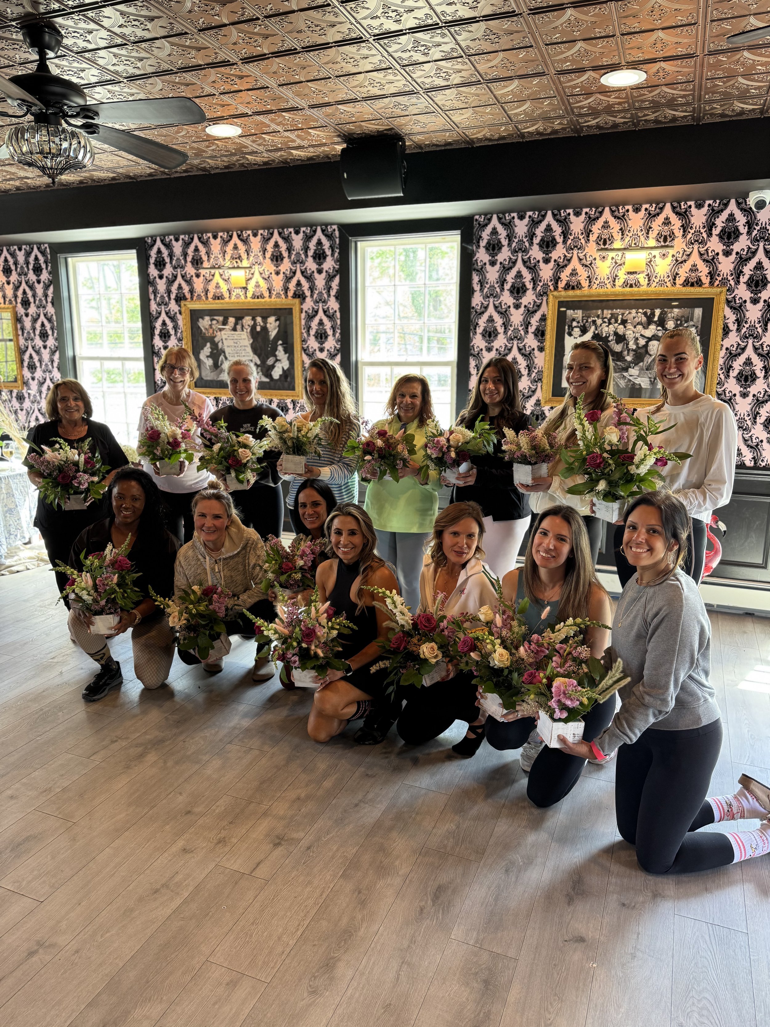 A group of women with flowers in pots posing for a photo in a decorated room with patterned wallpaper and large windows.