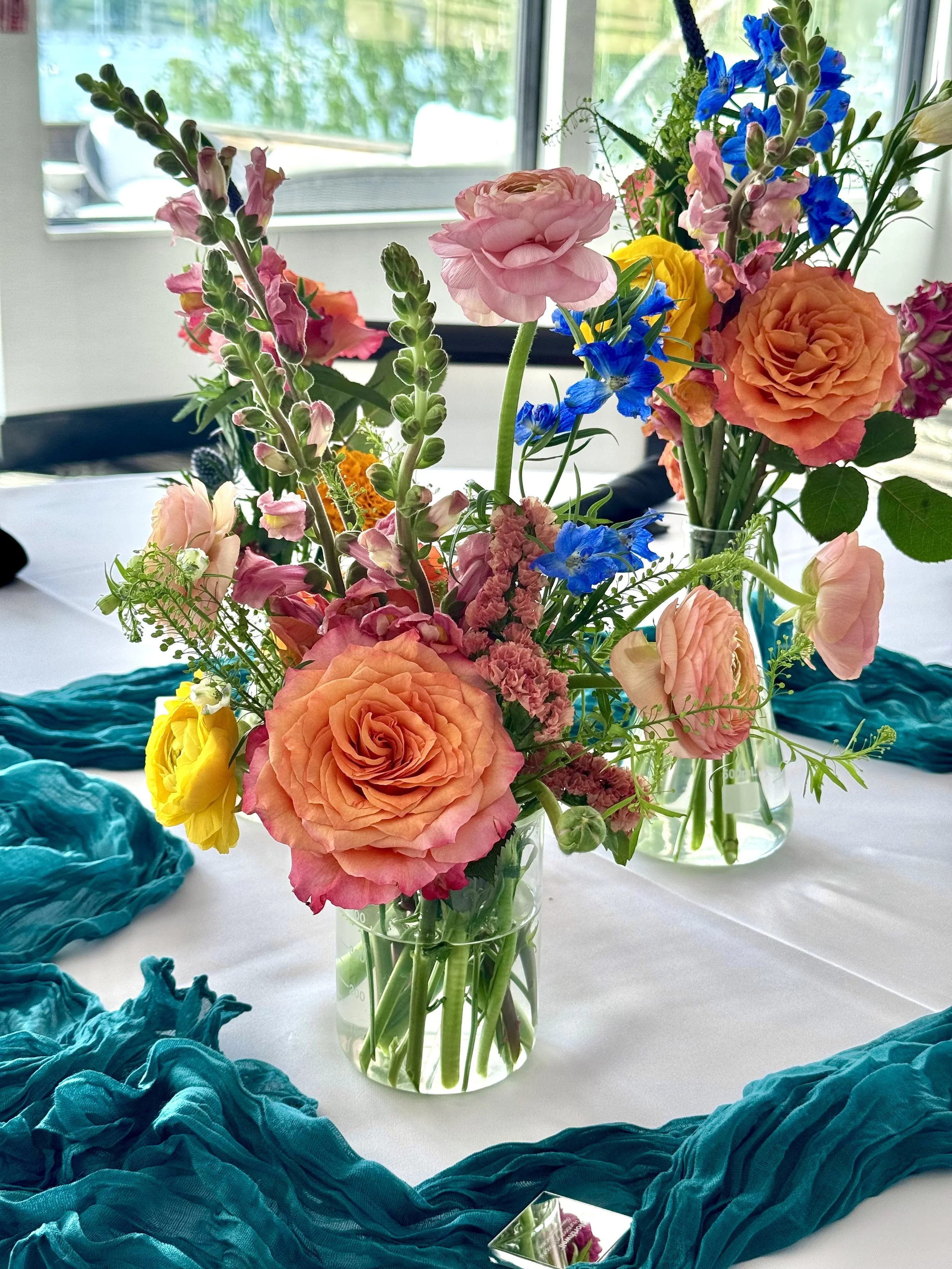 A table decorated with a glass vase filled with pink, peach, yellow, and blue flowers. Blue fabric is draped on the table surrounding the vase, and a mirror is partially visible in the foreground.