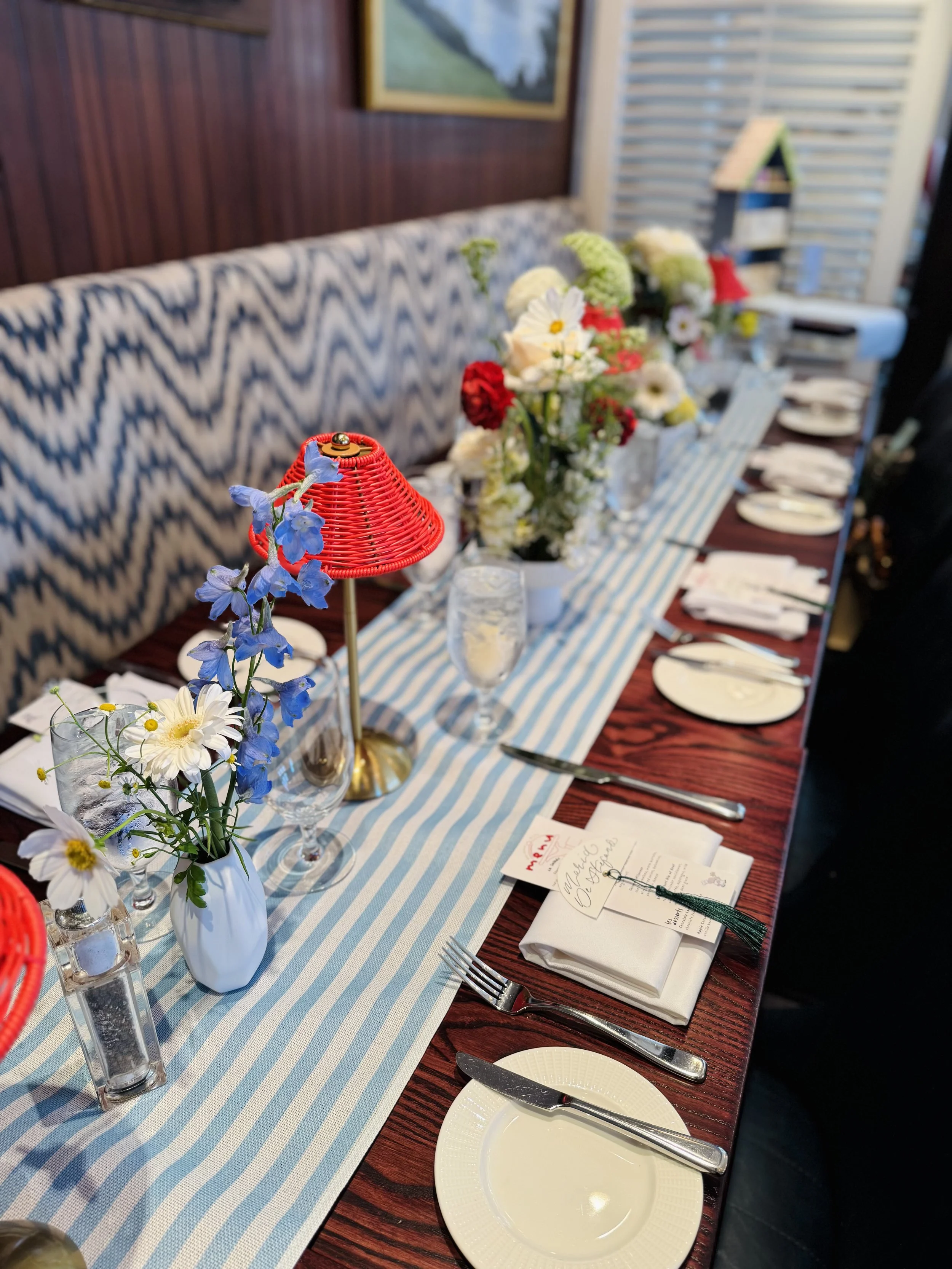 A long dining table with a blue and white striped table runner, decorated with vases of white, red, and blue flowers. There are white plates, napkins, and forks set at each place, and a small red lampshade with blue flowers on the table. A menu card and a pen are also on the table. The background includes a patterned bench, framed artwork, and a shelving unit with a small house-shaped decor piece.