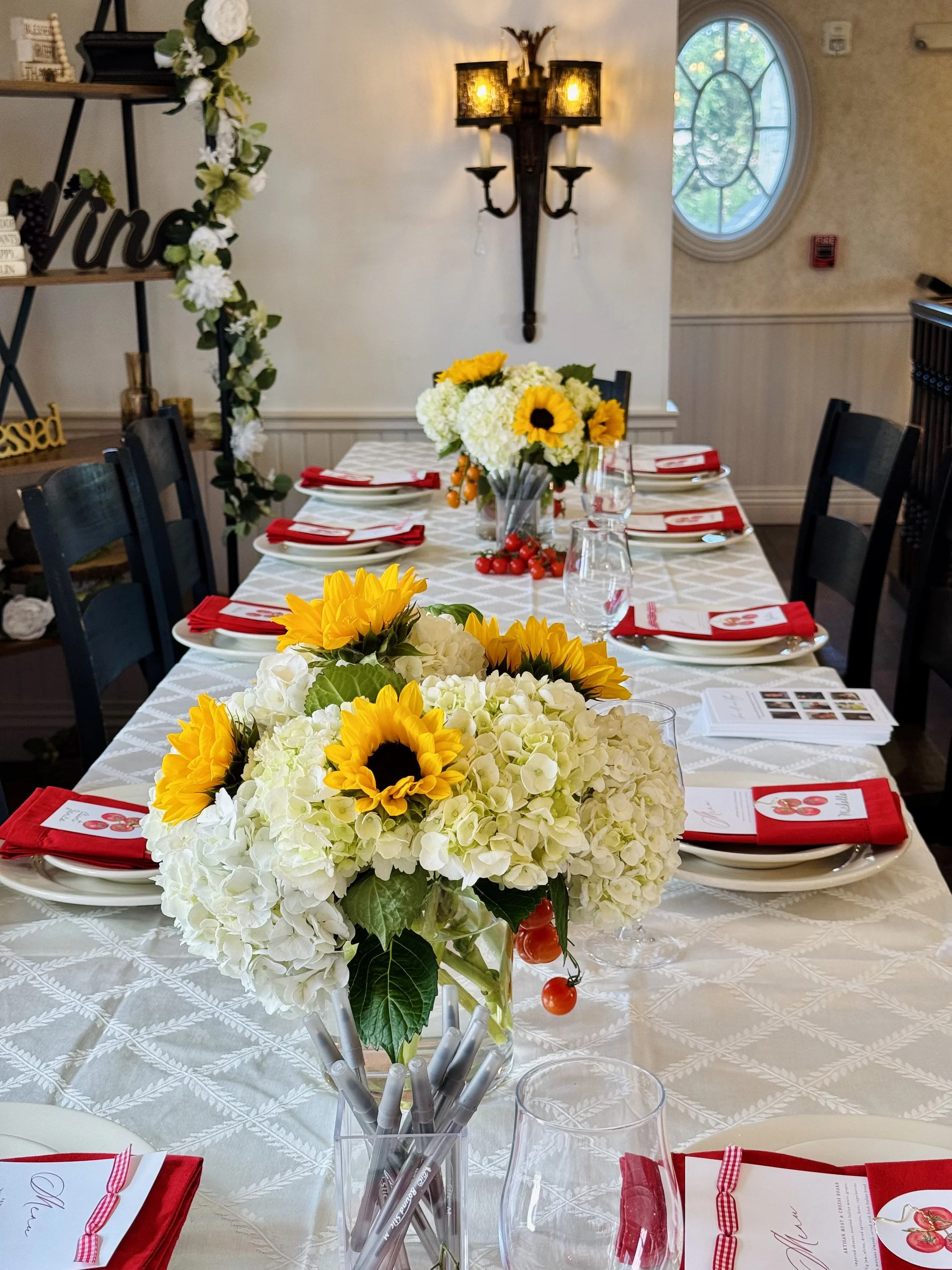 A decorated dining table set for an event with floral centerpieces, red napkins, and printed menus, in a room with a large window, floral wall decor, and warm lighting.