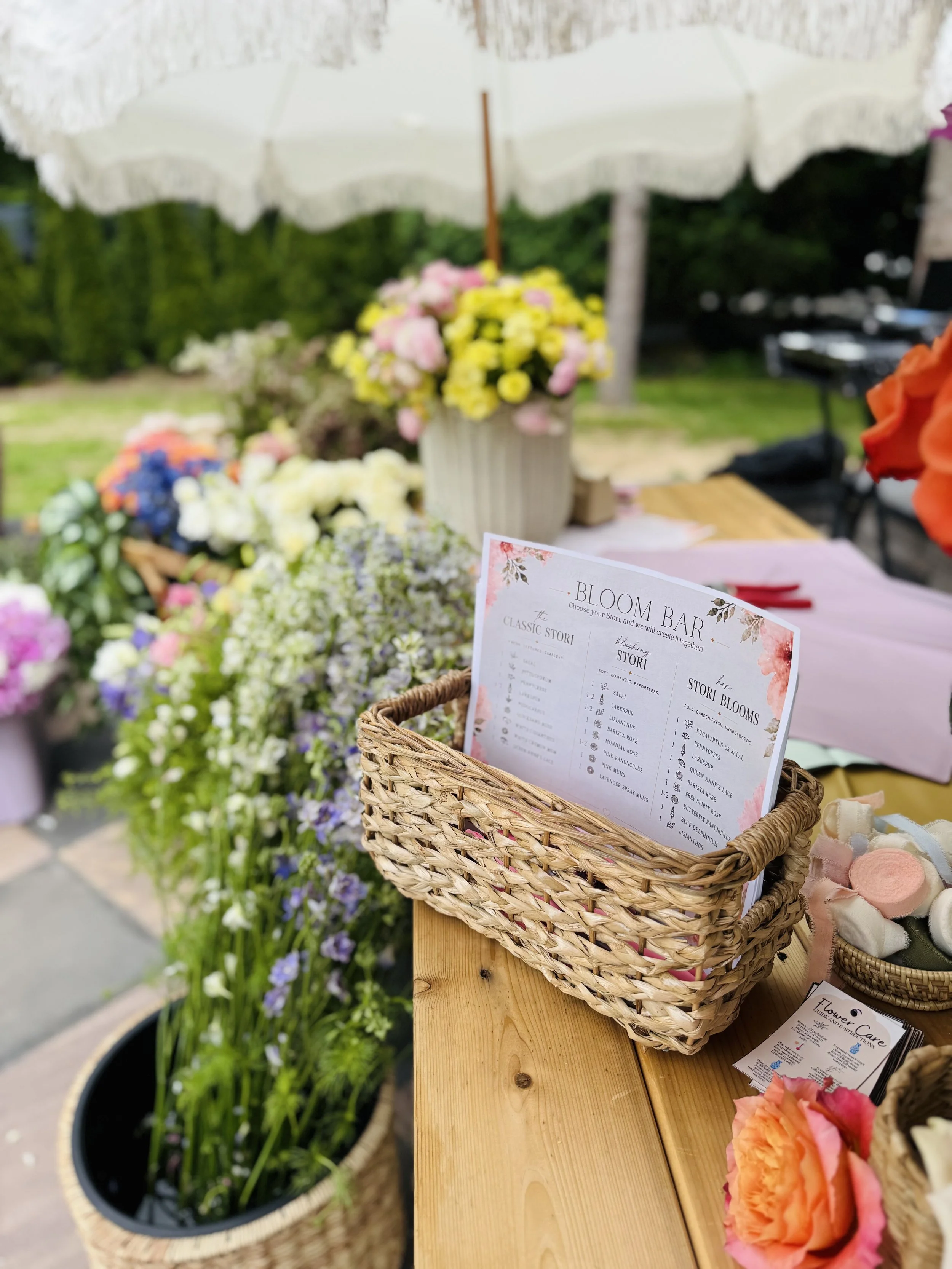 A display of colorful flowers, including pink, white, yellow, purple, and orange blooms, arranged in baskets and flower pots on a wooden table at an outdoor market or garden event.