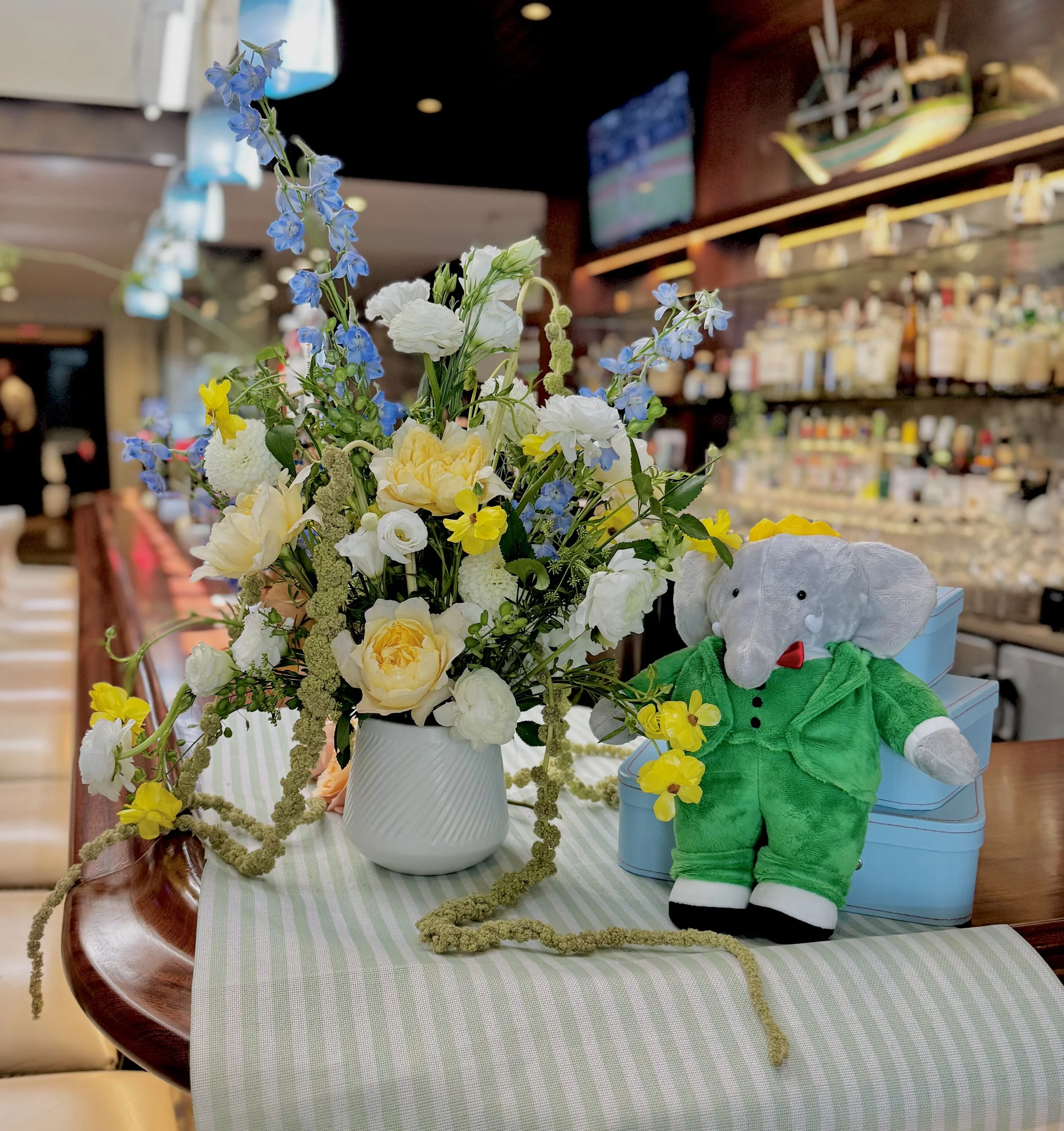 A table with a green and white checkered tablecloth holding a white vase with a bouquet of yellow, white, and blue flowers, and a white stuffed animal dressed in a green and red outfit. The background features shelves with bottles and a television sc
