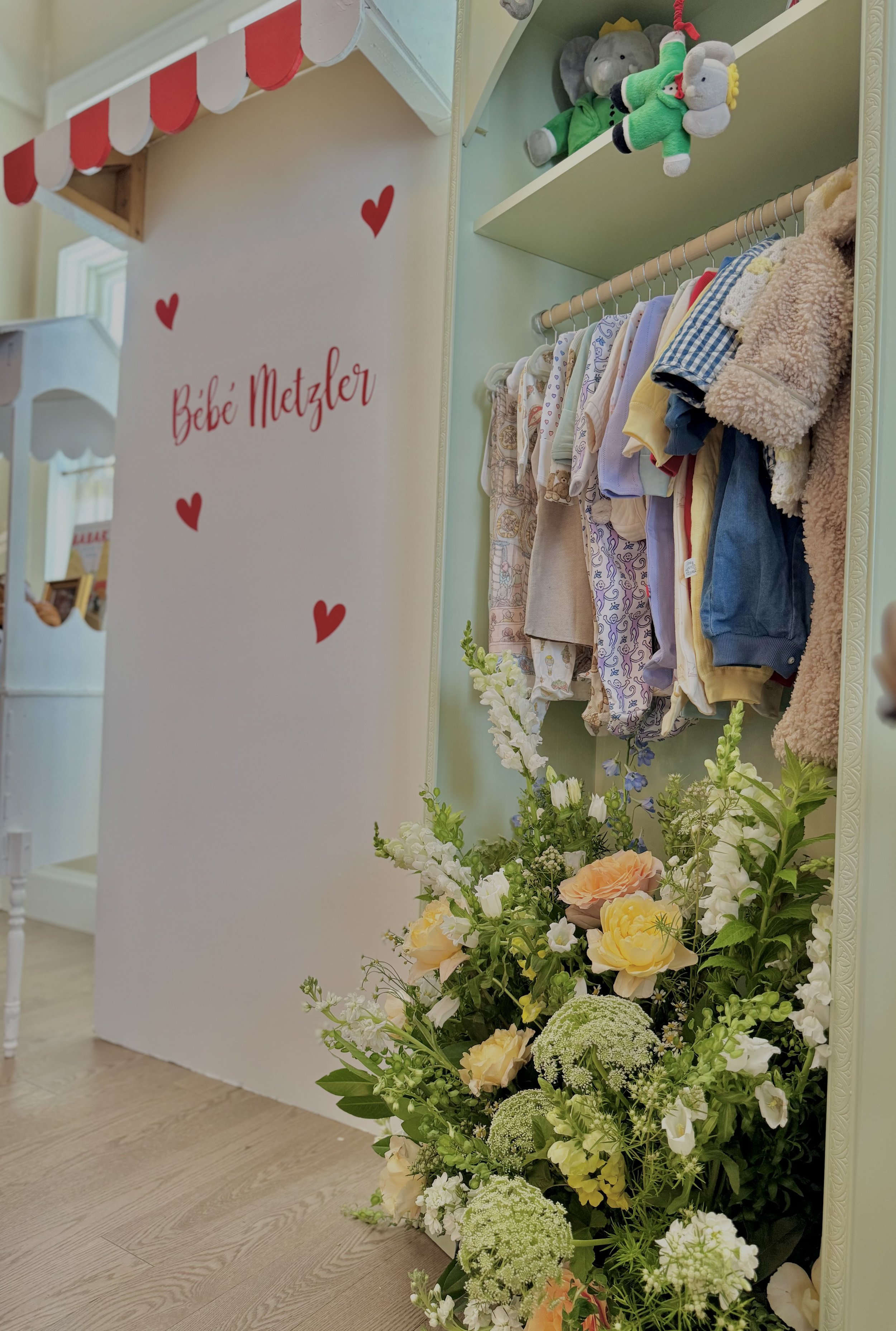 A closet filled with baby clothes on a rack, plush toys on a shelf, and a large floral arrangement in front, with a white wall that has red hearts and the words "Babe Metzler" written on it.
