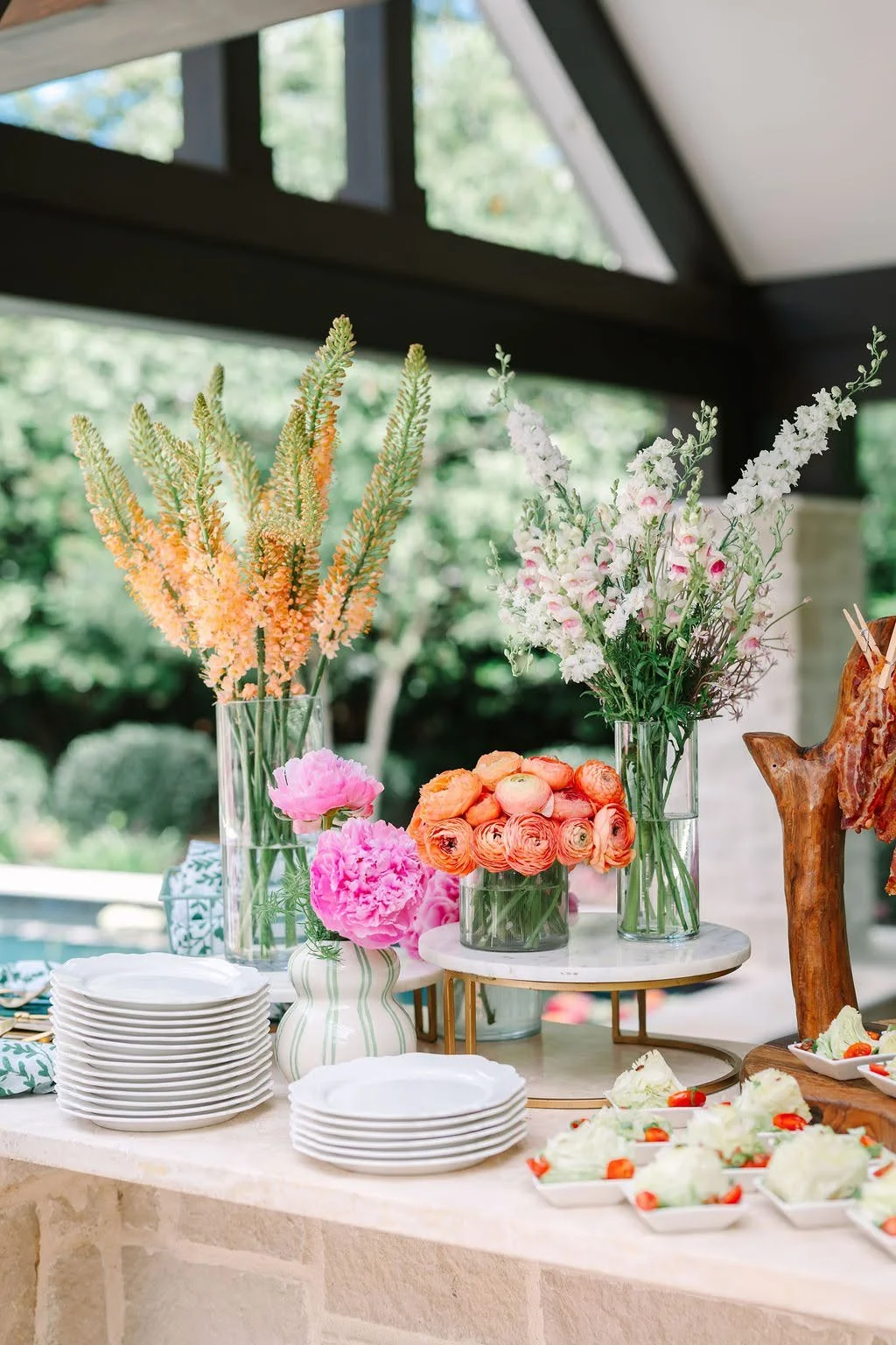 Gilded Event & Floral Design — Decorative table with colorful floral arrangements, white plates, and small appetizers in a bright, indoor setting with greenery visible outside.
