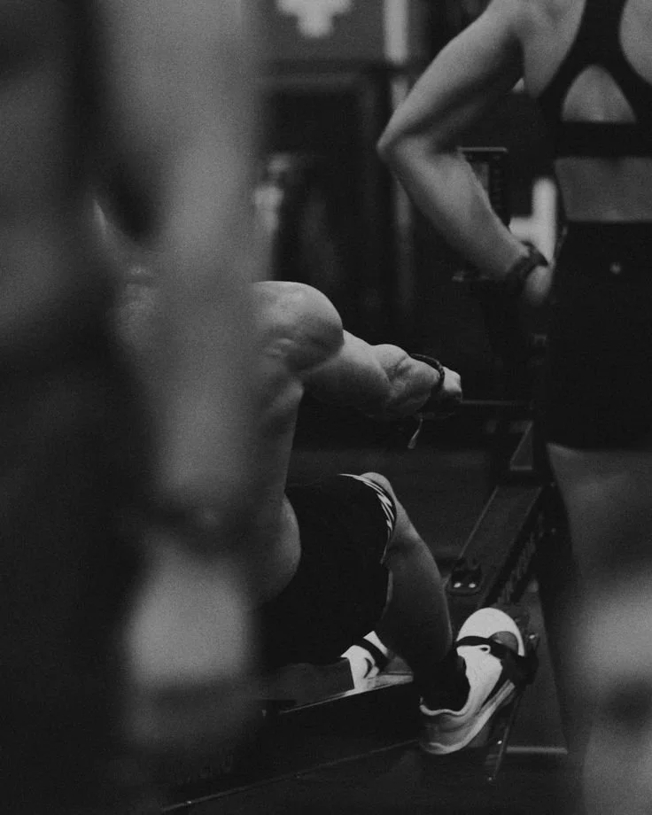 A person in workout attire on a rowing machine in a gym, with another person standing nearby.