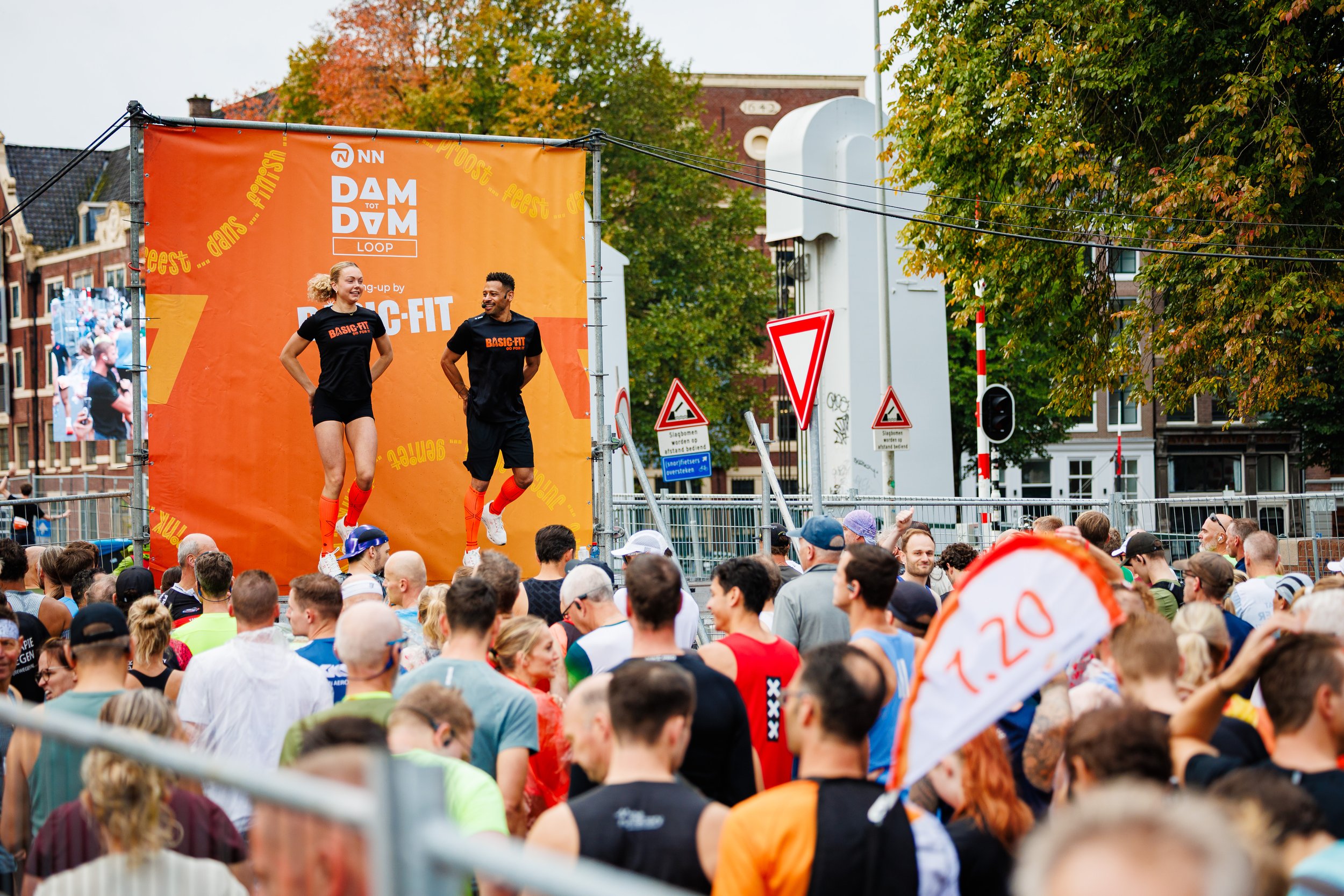 Two performers in black shirts and shorts jump on a stage with an orange backdrop during an outdoor event. A large crowd watches them, and colorful buildings and trees are in the background.