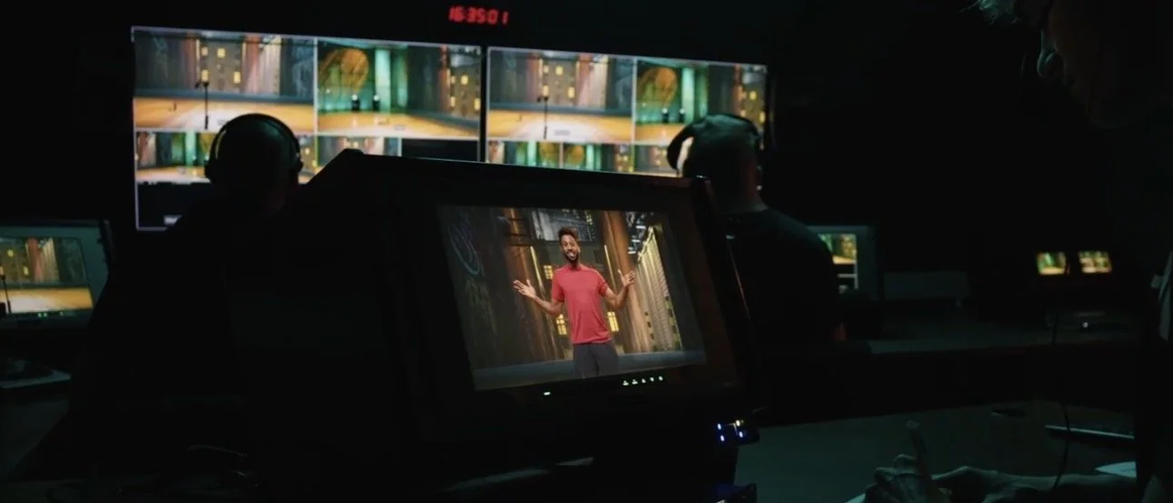 A video production control room with multiple monitors, showing a man in a red shirt speaking on stage during a recorded or live event.