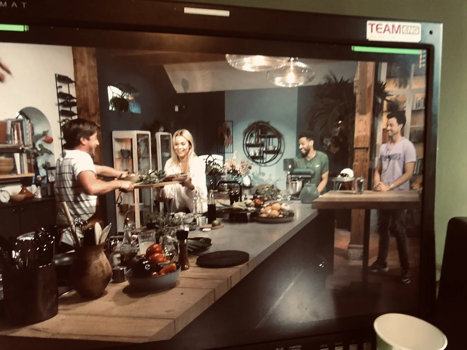 People preparing food in a kitchen seen through a glass window, with four people visible, including two men and two women, smiling and working together amid kitchen appliances, dishes, and food ingredients.