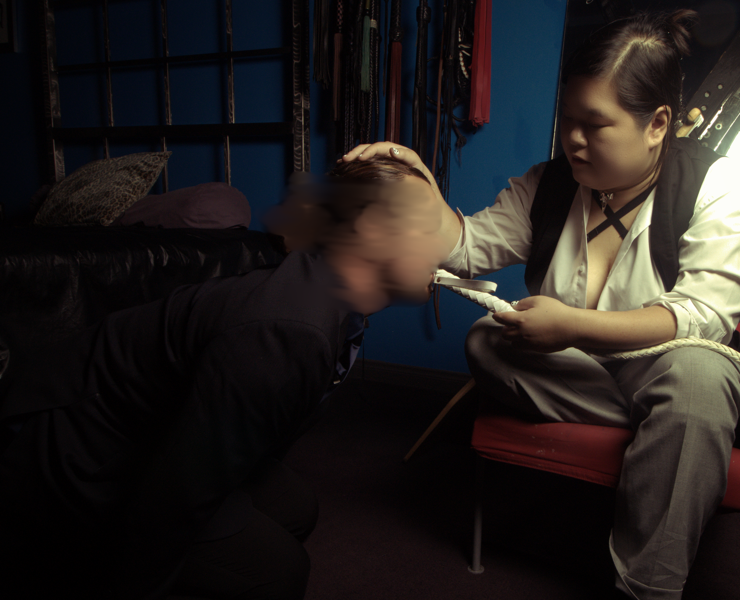 Goddess Shi (Goddess Shiney), an Asian dominatrix, wears a white blouse and black vest, and is holding out the end of a white whip to a male submissive in a black suit. The submissive kisses the end of the whip, while kneeling at her feet.
