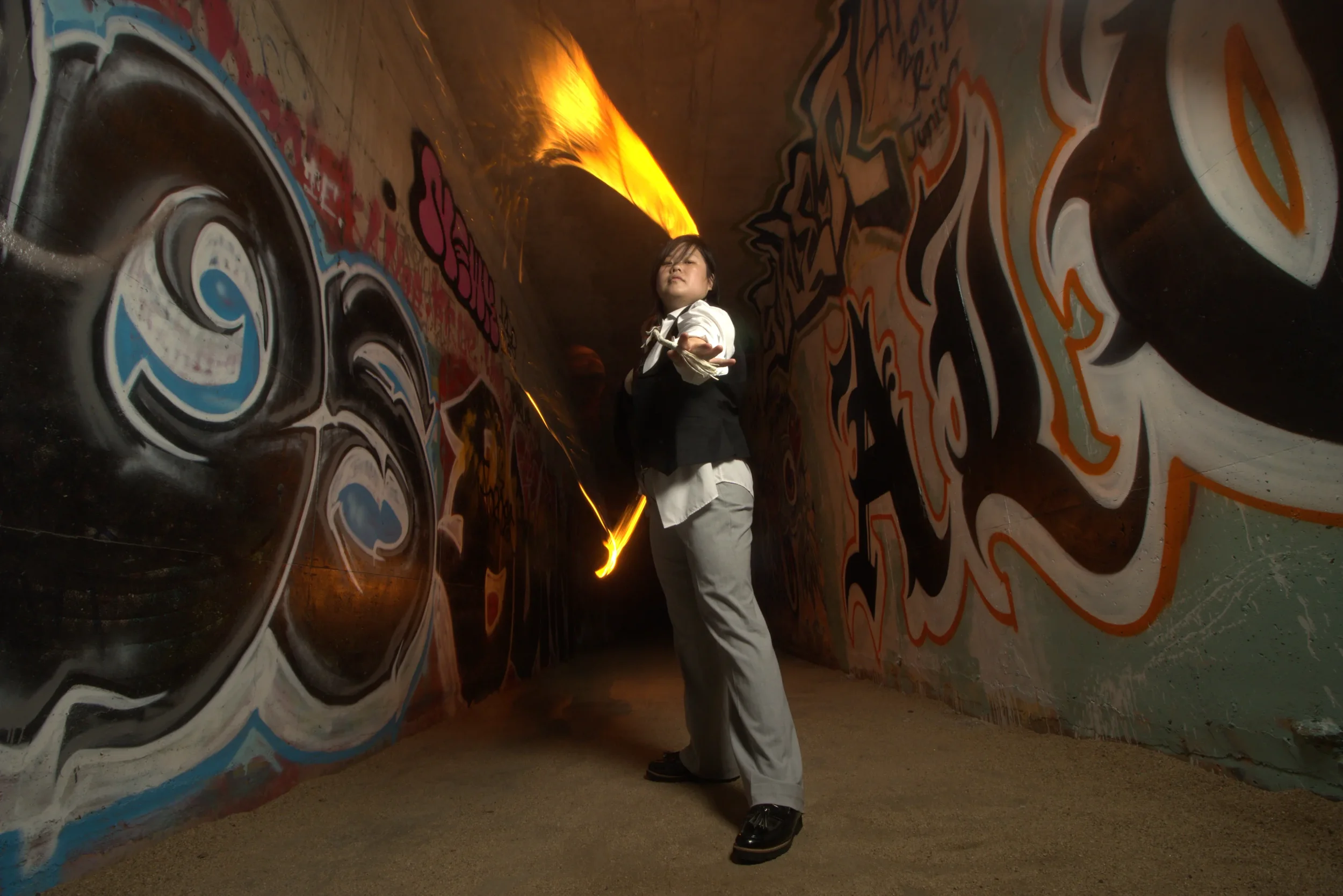 Goddess Shi (Goddess Shiney) in formal attire in a graffiti-covered tunnel, fire streaking behind her.