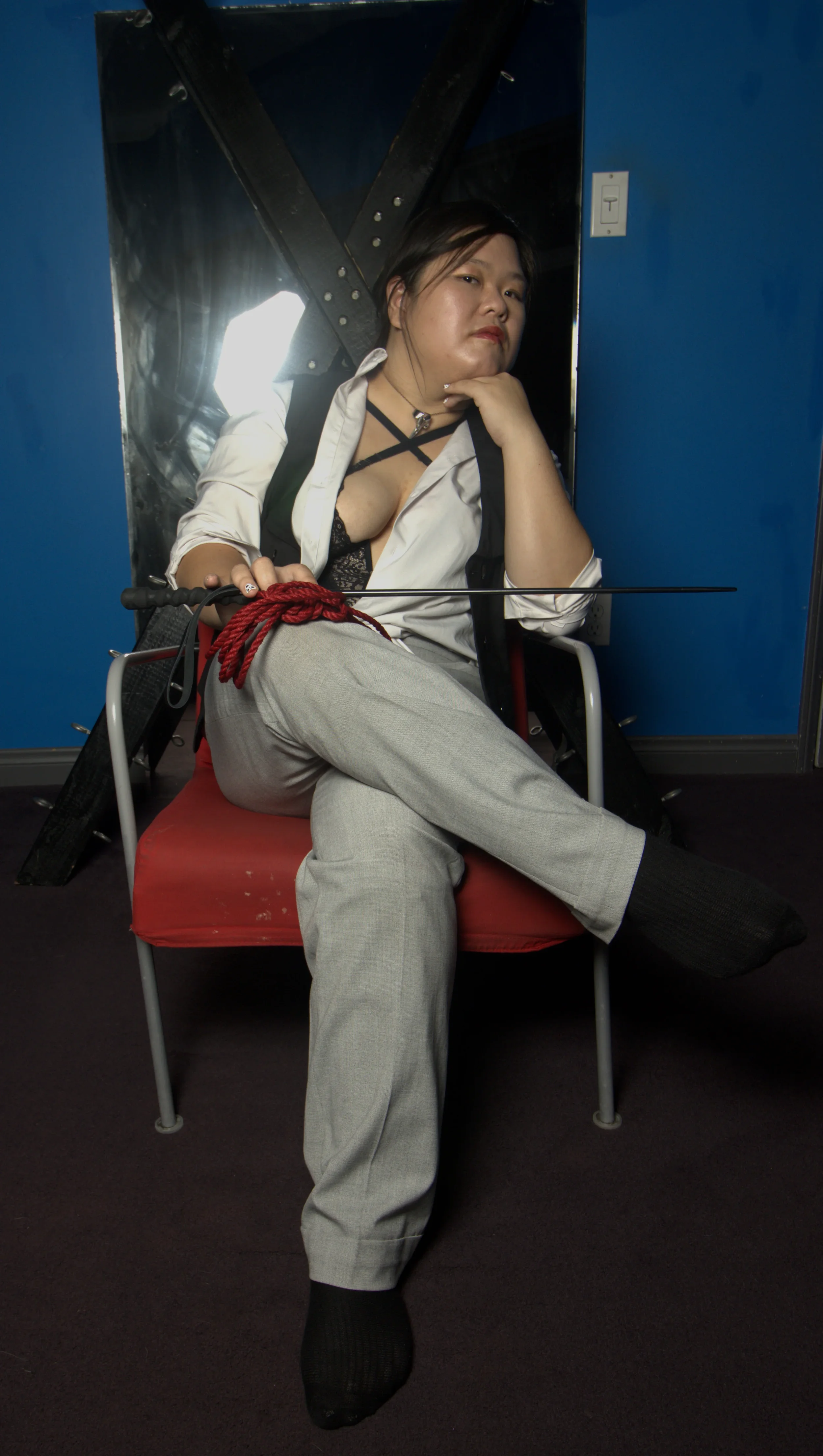 Goddess Shi (Goddess Shiney), an asian dominatrix, sits legs crossed on a red seat in front of a St Andrew's Cross and a mirror. She has red rope in her lap and a black switch. She side eyes the camera, thinking of plans and schemes.