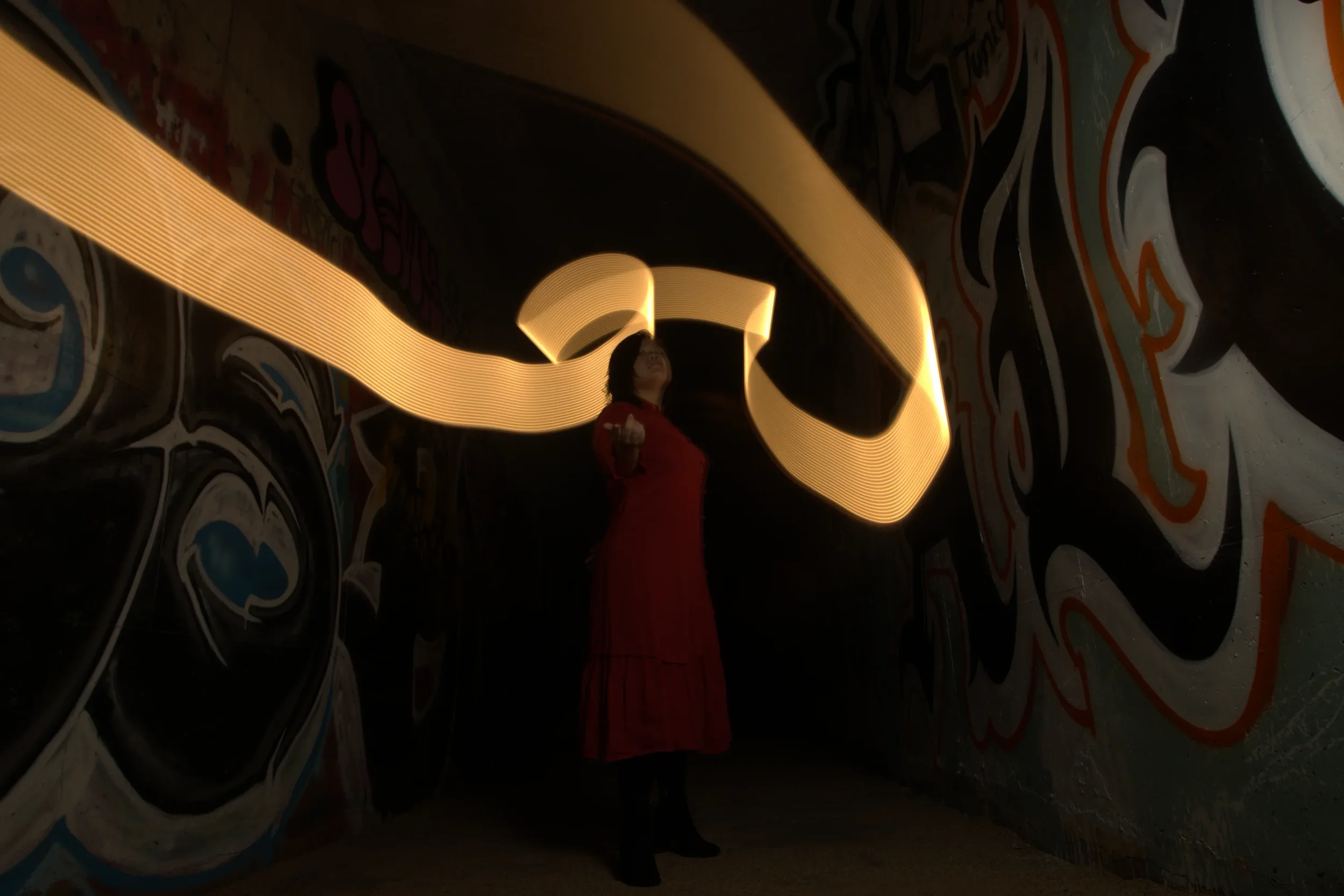 Goddess Shi in a red dress standing in a graffiti-covered tunnel, holding a light trail creating a glowing swirl above her head. She holds out her hand, beckoning the viewer.