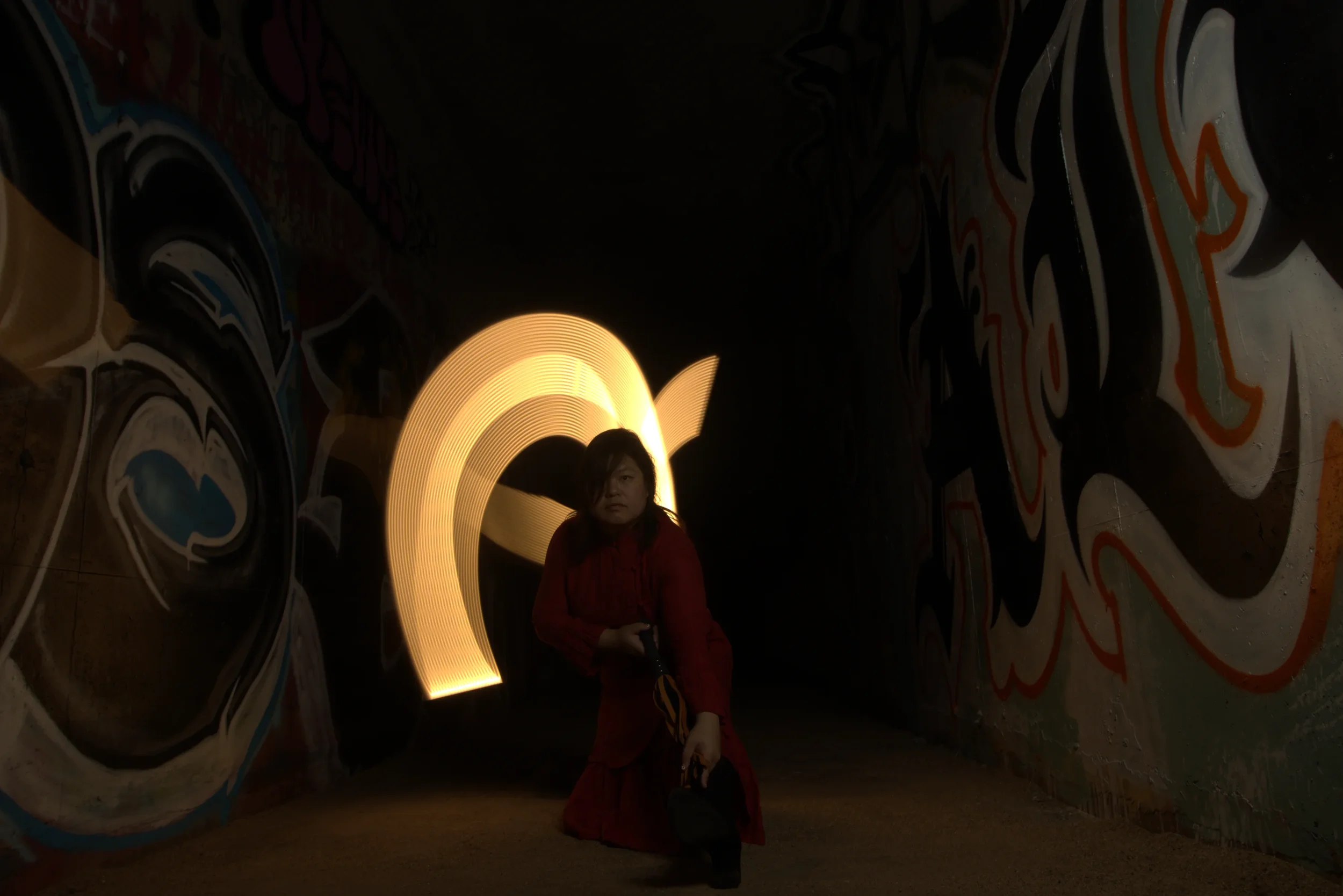 Goddess Shi (Goddess Shiney) in a red dress crouches in a dark alley with mural-covered walls, illuminated by a large, glowing, crescent-shaped light.