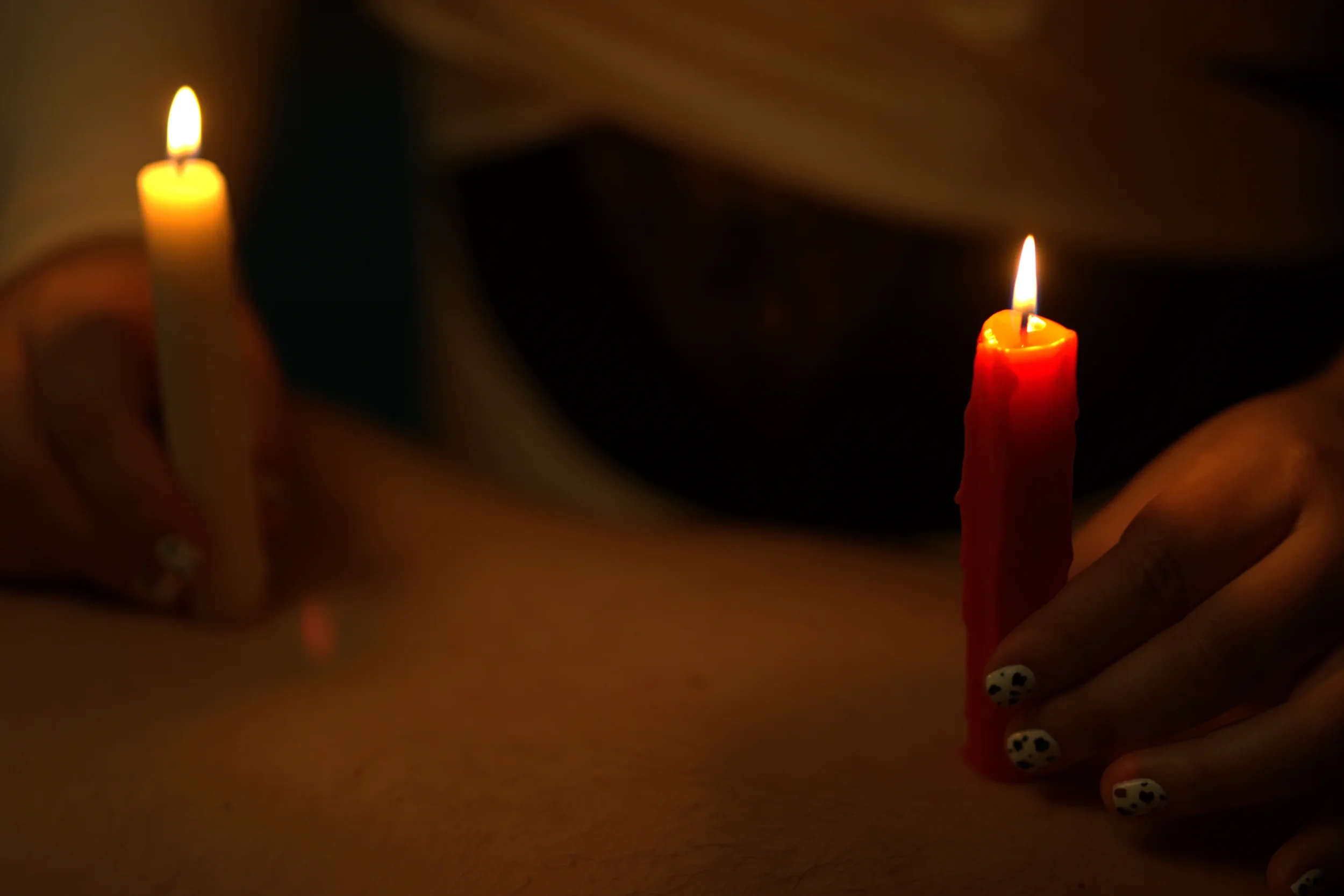 Goddess Shi lights two candles on the back of her submissive, allowing the wax to melt onto their skin. The scene is dim except for the flames.
