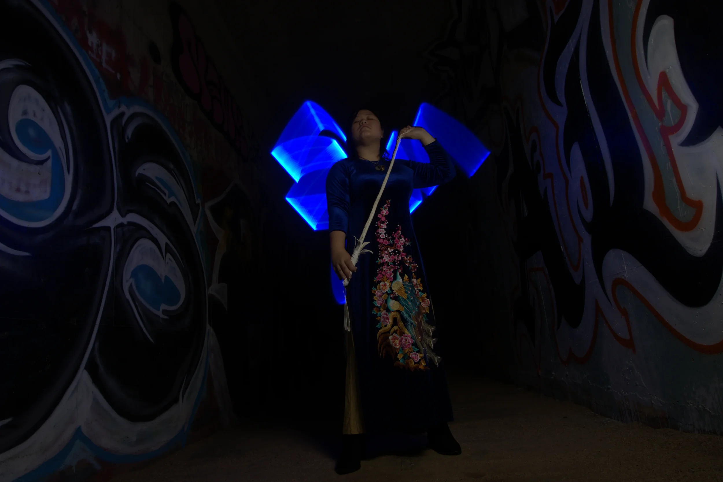 Goddess Shi (Goddess Shiney) wearing a traditional embroidered dress holds a whip in a dimly lit space with graffiti on the walls, illuminated by blue neon lights and large glowing electric blue wings behind her.