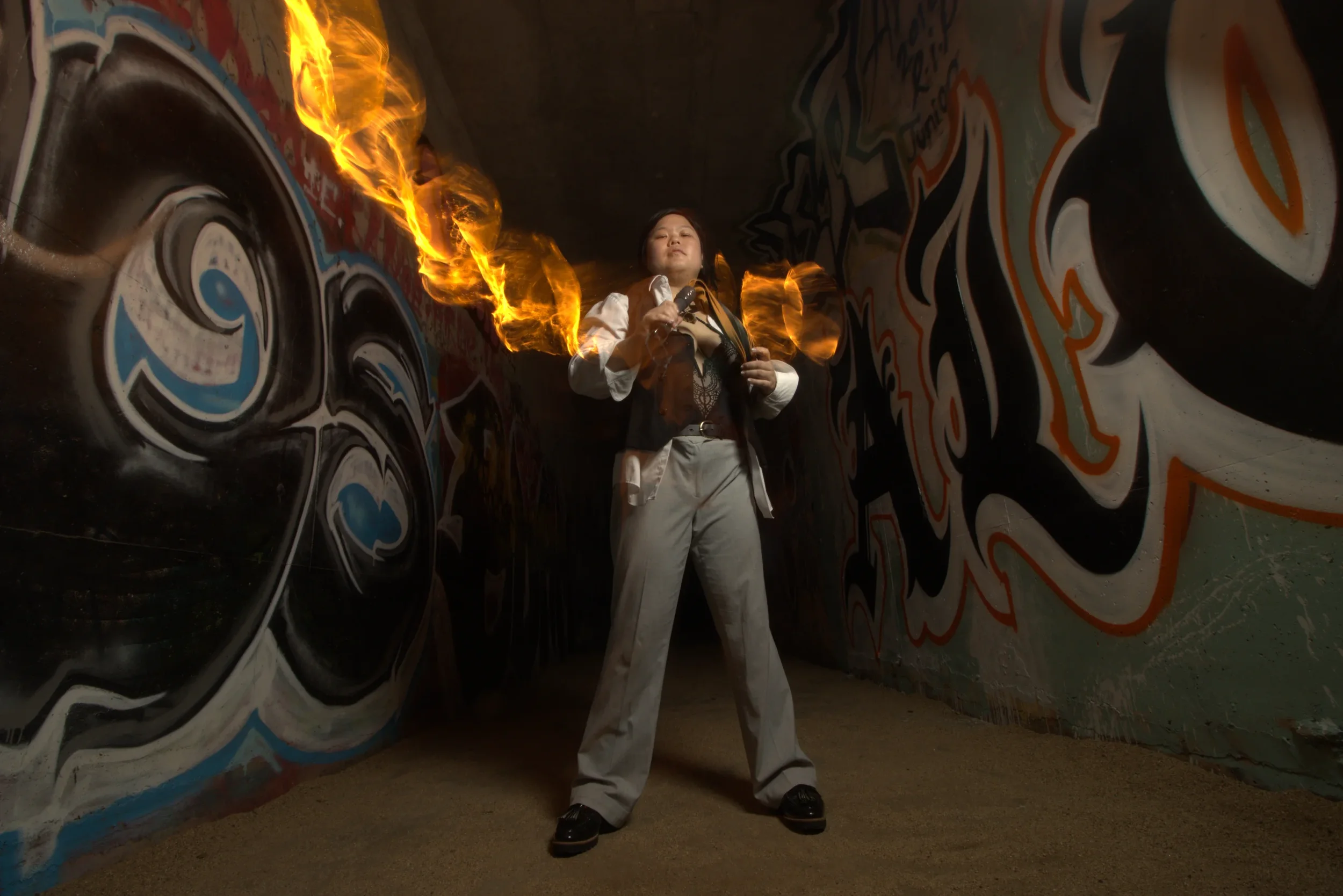 Goddess Shi (Goddess Shiney) stands in a graffiti-covered tunnel holding a tool with flames coming from behind them.