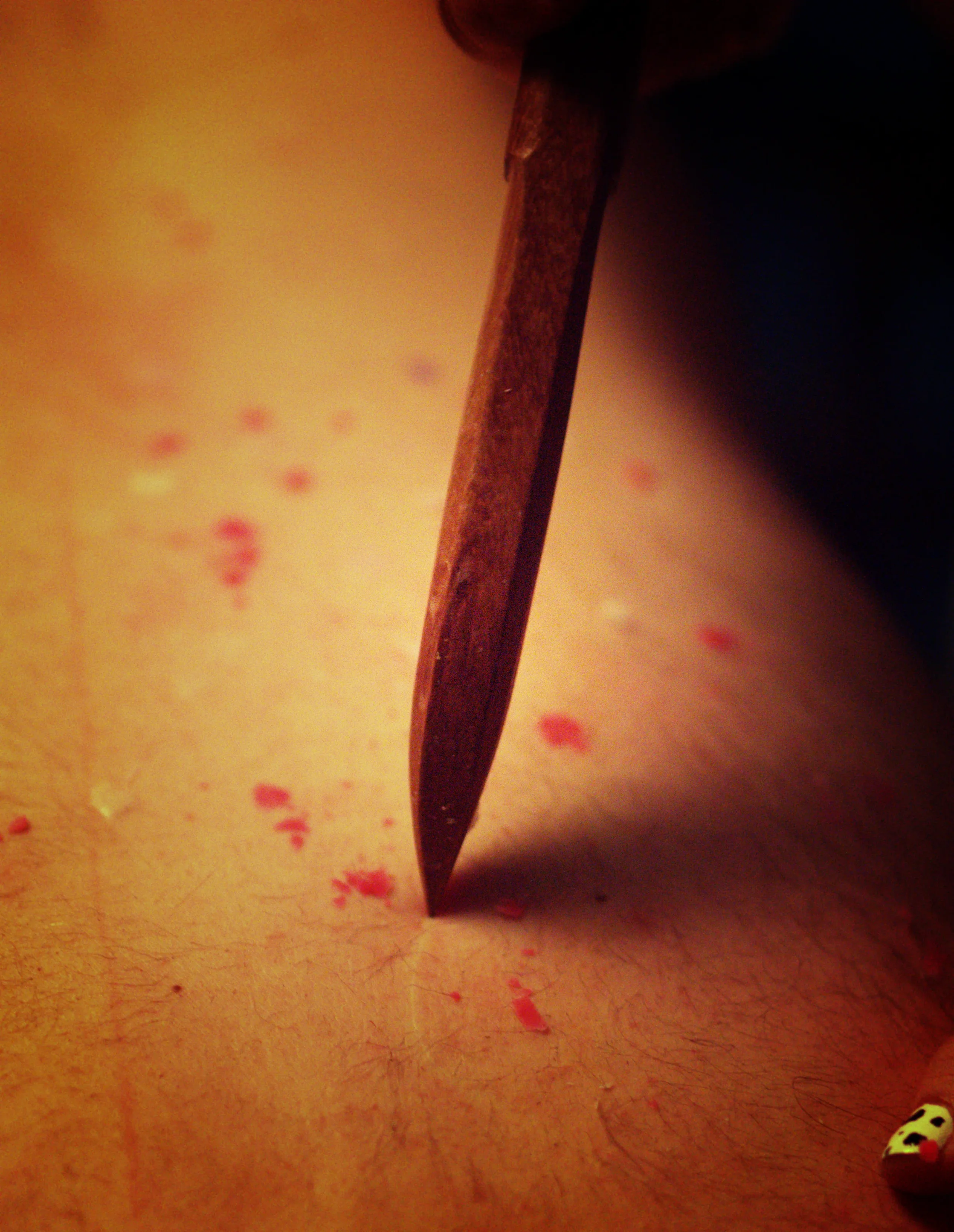 A wooden knife scraping against the skin, with splatters of red and white wax dotted around. A white line follows the trail of the knife.