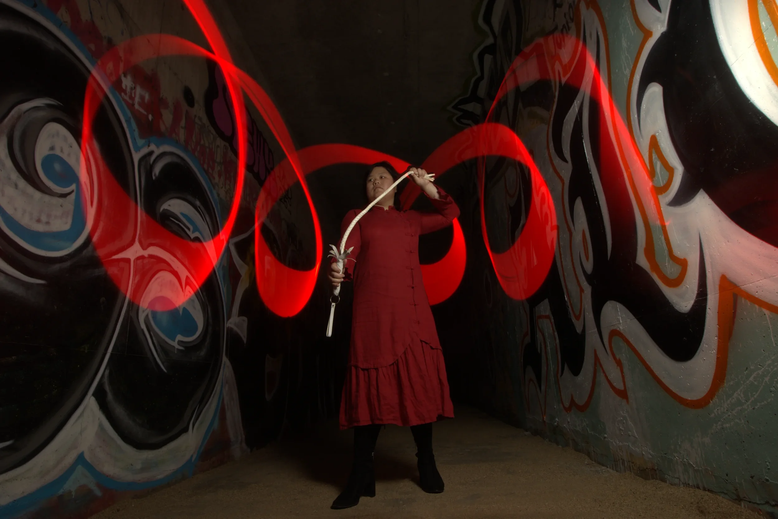 Goddess Shi (Goddess Shiney) in a red dress holding a whip in a tunnel with graffiti on the walls, illuminated by red light trails.