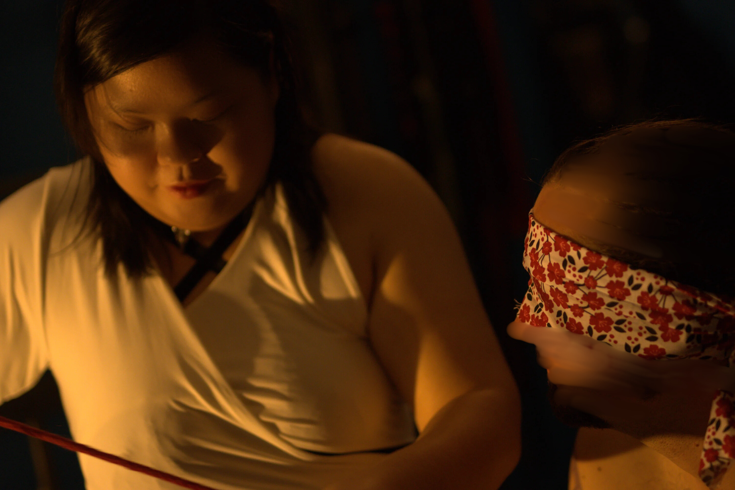 Goddess Shi (Goddess Shiney), an Asian dominatrix, wears a white dress, and prepares a red rope while her male submissive is blindfolded with a red floral cloth. He seems to look in her direction.