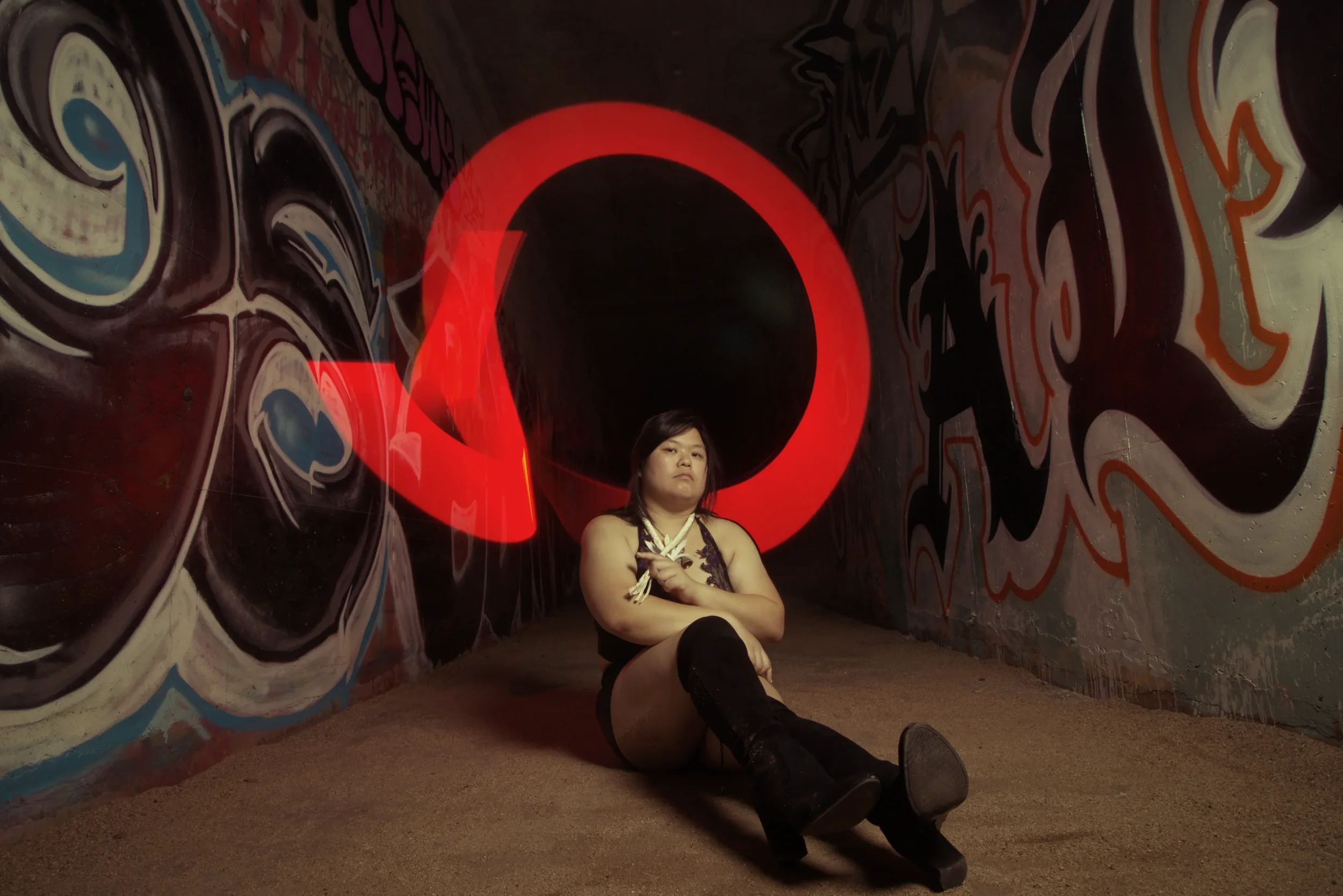 Goddess Shi (Goddess Shiney) sitting on the ground in a graffiti-covered tunnel with a large red glow behind her.