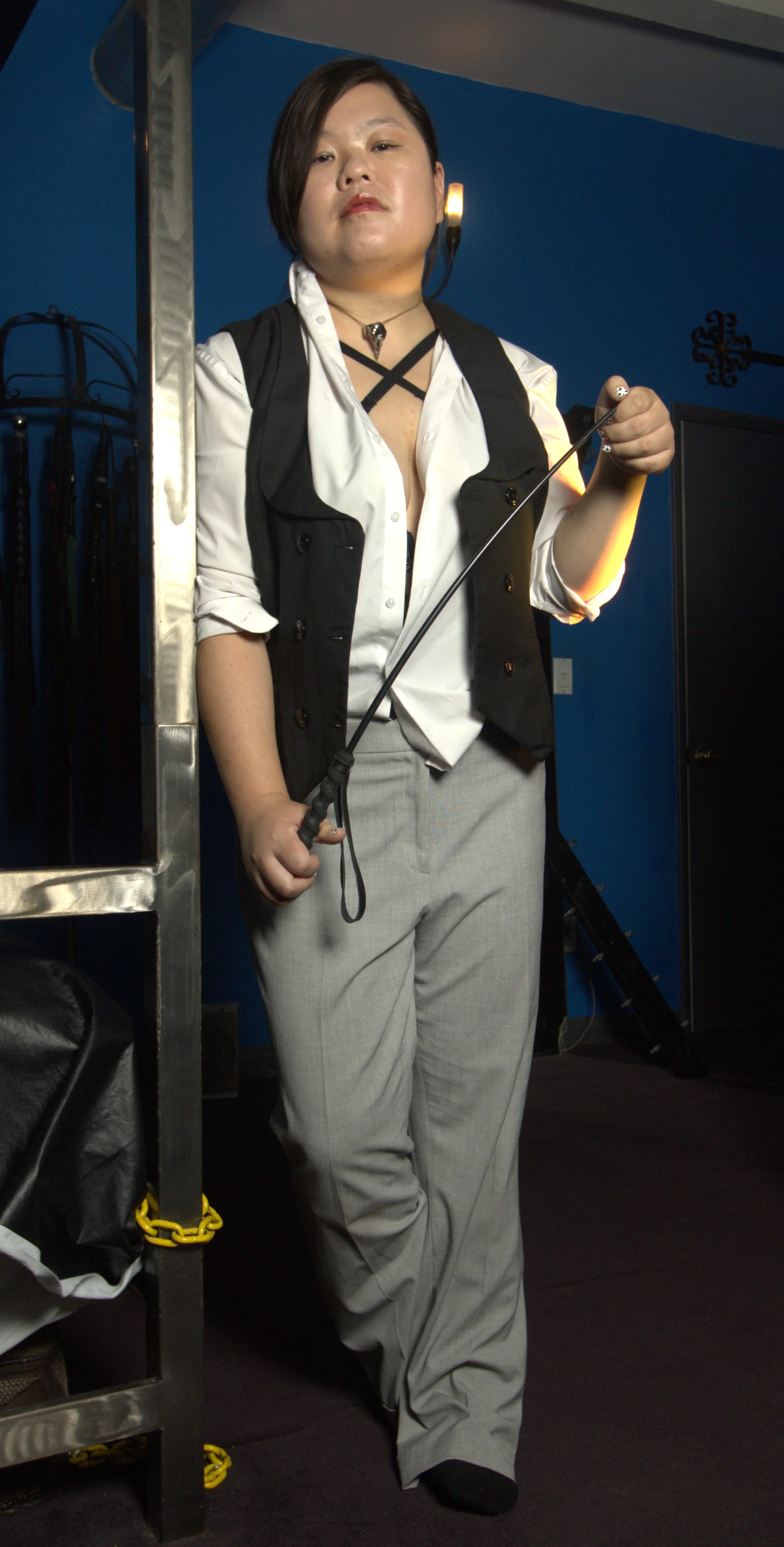 Goddess Shi (Goddess Shiney), an Asian dominatrix, leans against a metal post in business casual wear, holding a crop, and smiling.