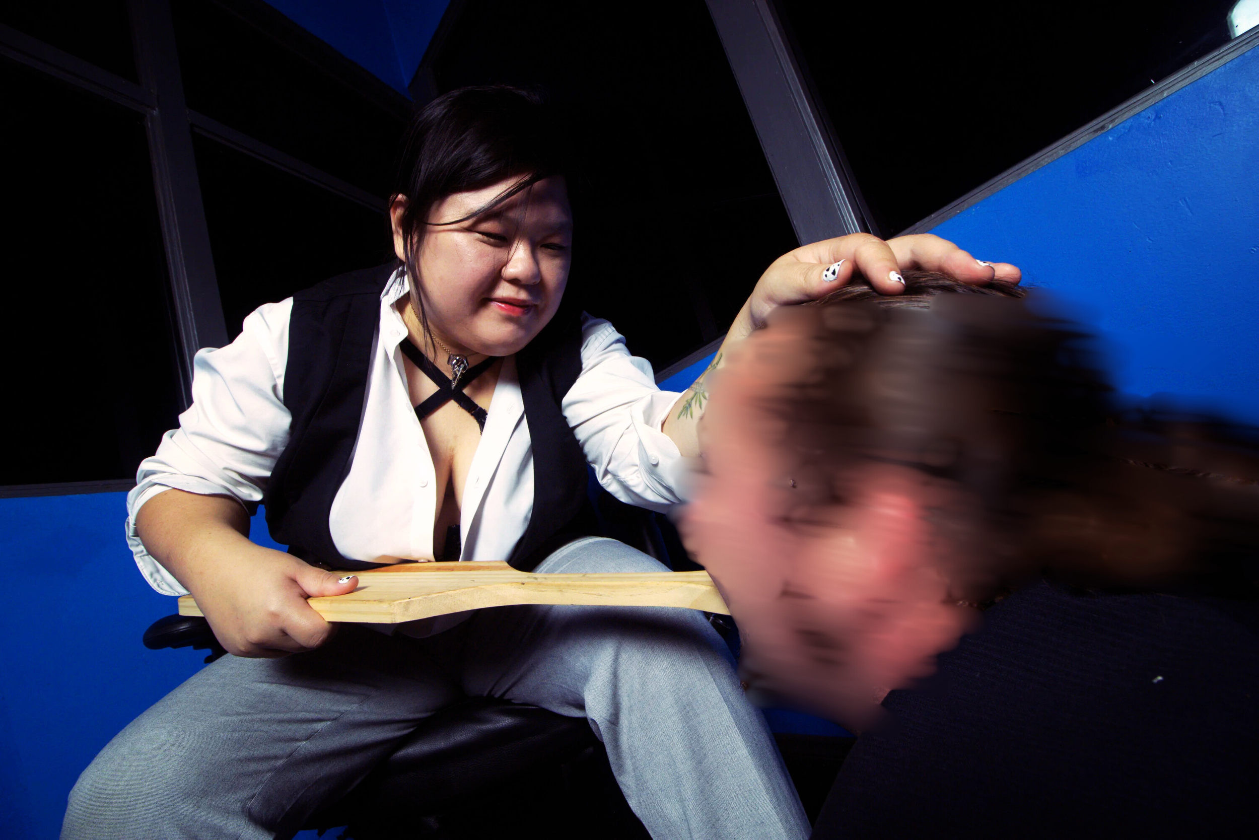 Goddess Shi (Goddess Shiney), an Asian dominatrix, wears a white blouse and black vest, is holds her male submissive's head. He looks up at her, kissing her paddle. She smiles gently at him, pleased.