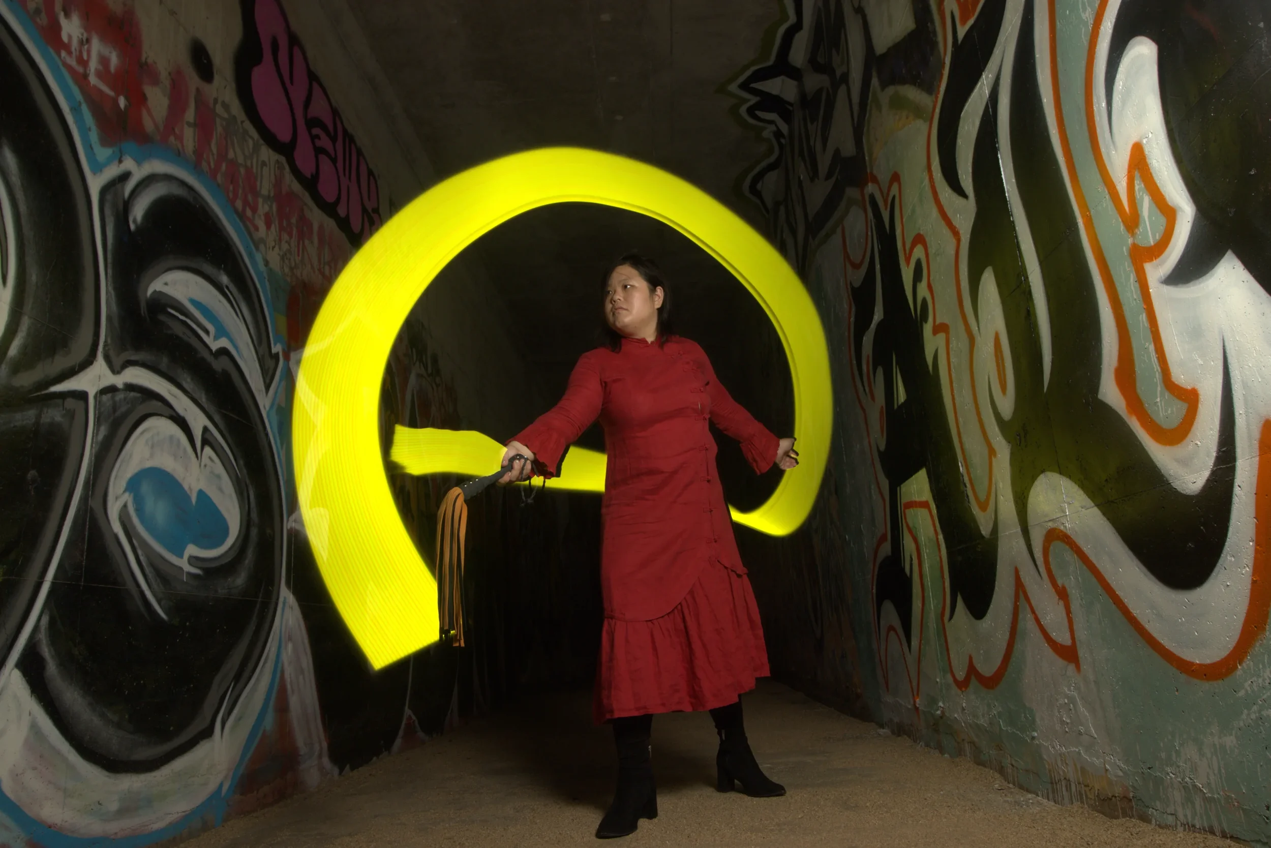 Goddess Shi (Goddess Shiney) in a red dress and black heeled boots stands in a graffiti-covered tunnel with a glowing yellow light sculpture shaped like a question mark behind her.