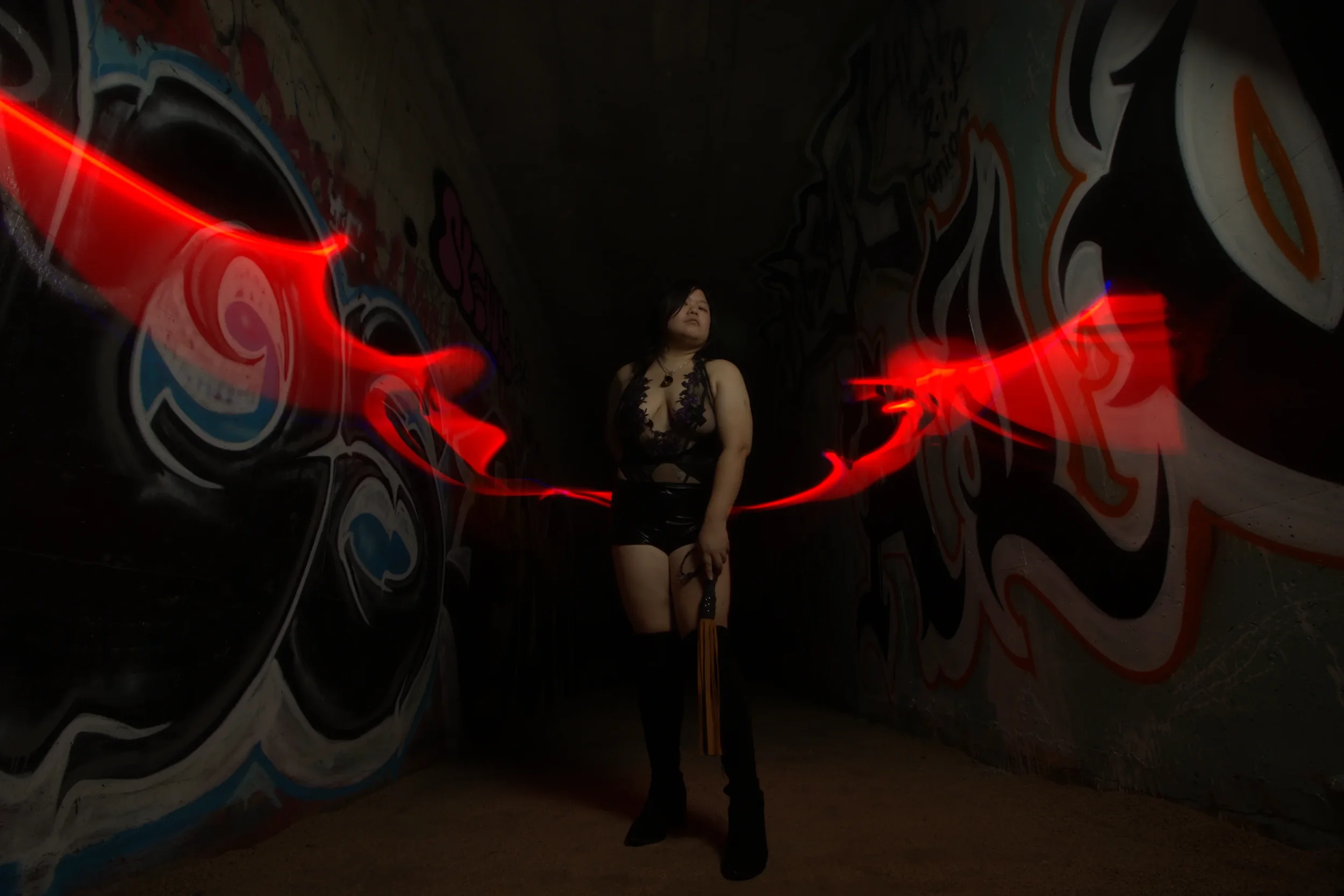 Goddess Shi (Goddess Shiney) standing in a graffiti-covered tunnel surrounded by neon red light trails, wearing a black lace top and high boots. She has her flogger in front of her.