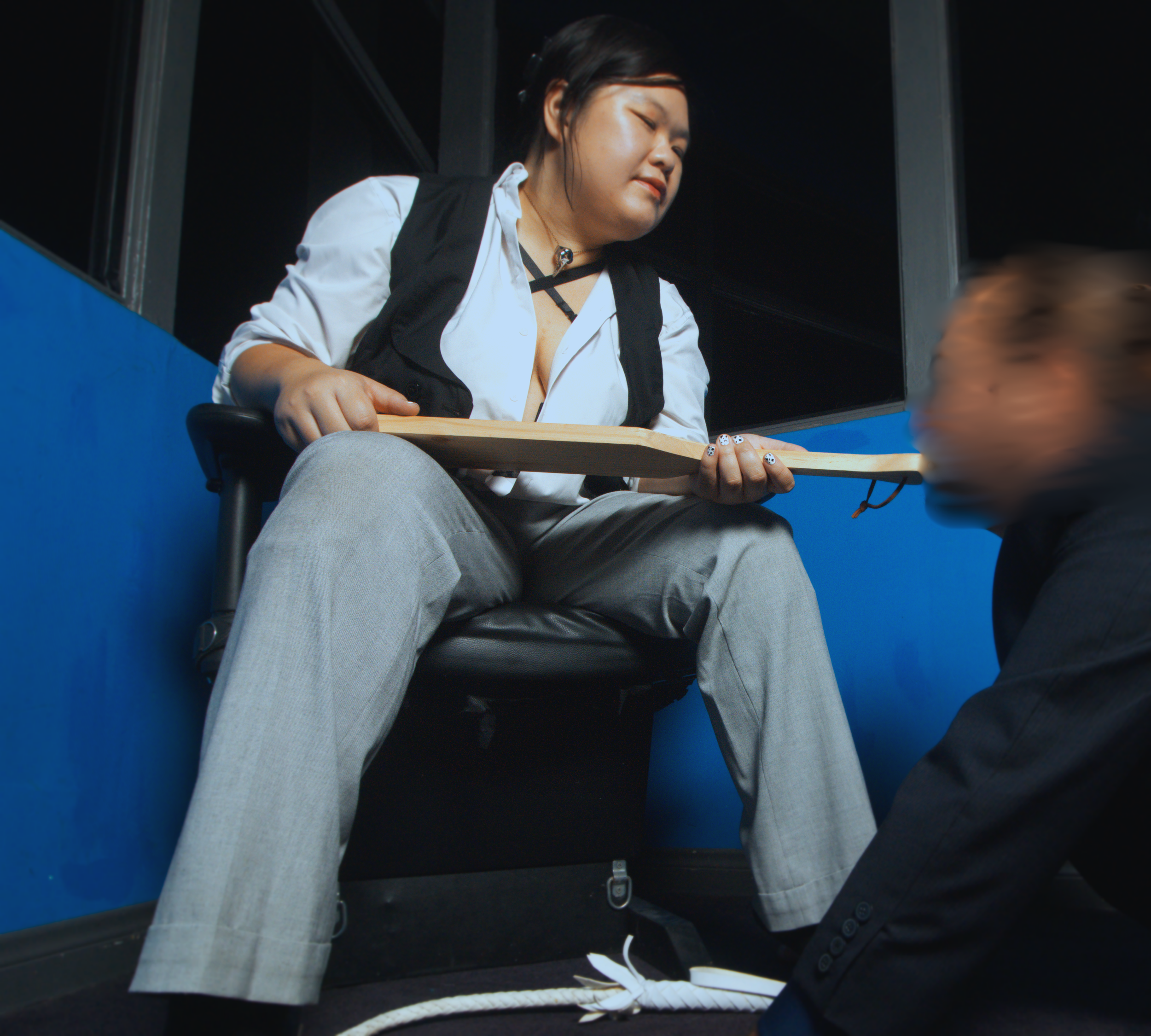 Goddess Shi (Goddess Shiney), an Asian dominatrix, wears a white blouse and black vest, holds her paddle out to a male submissive in a black suit, knelt at her feet. The male submissive kisses the end of the paddle.