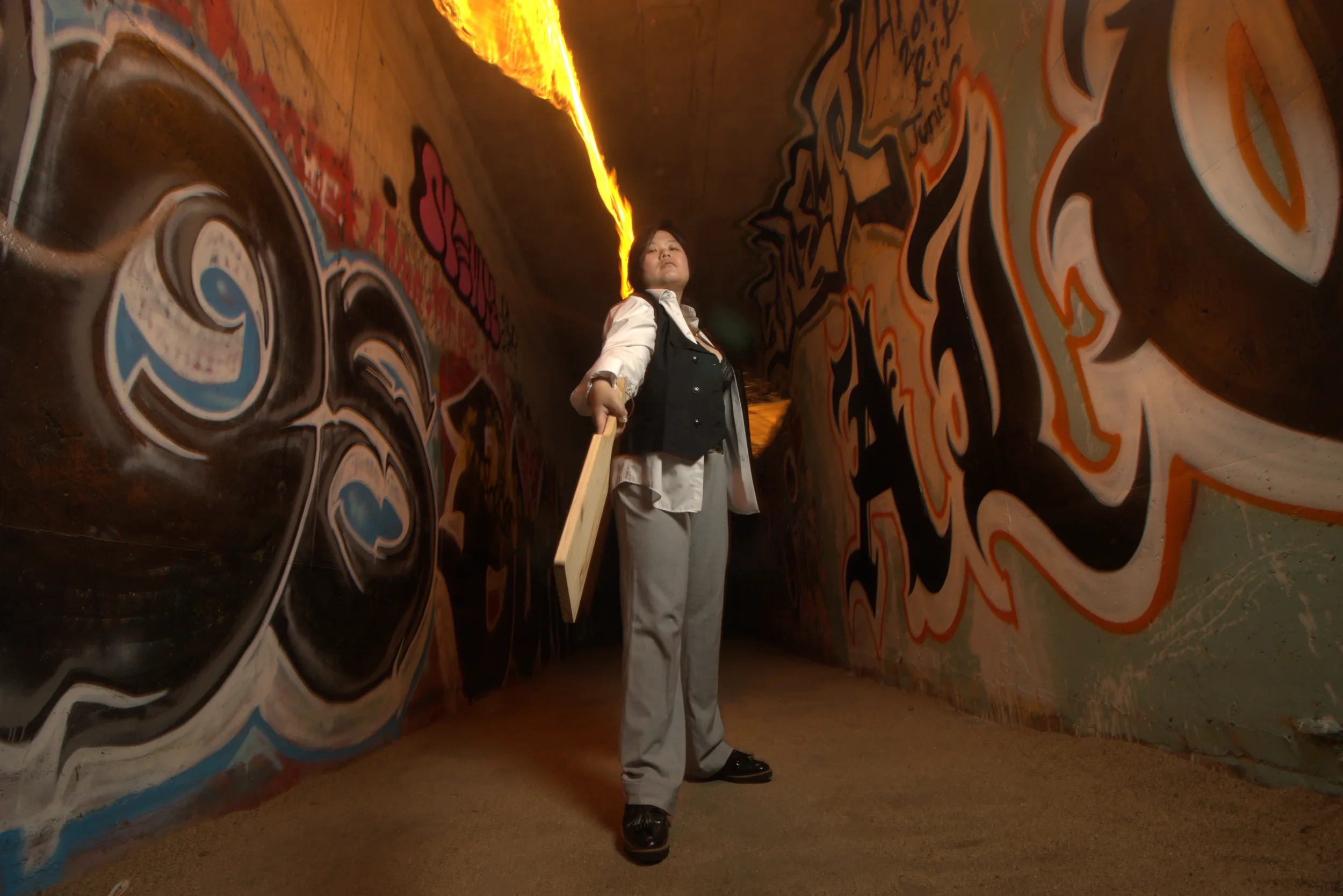 Goddess Shi (Goddess Shiney) in a suit holding a wooden plank stands in a graffiti-covered tunnel with vibrant wall art and a glowing fire above.