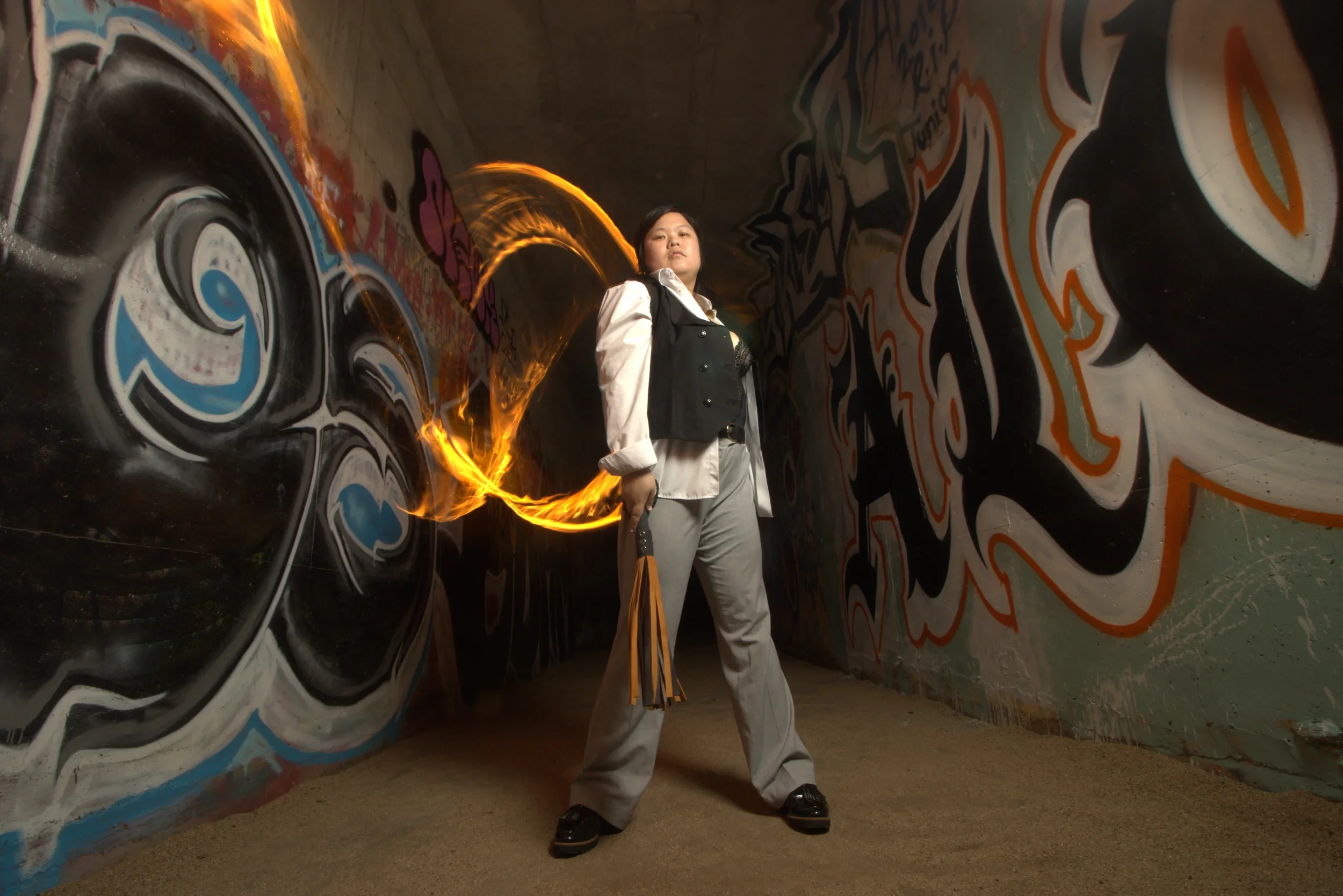 Goddess Shi (Goddess Shiney) standing in a graffiti-covered tunnel, holding a flaming torch with fire swirling behind her, holding her flogger.