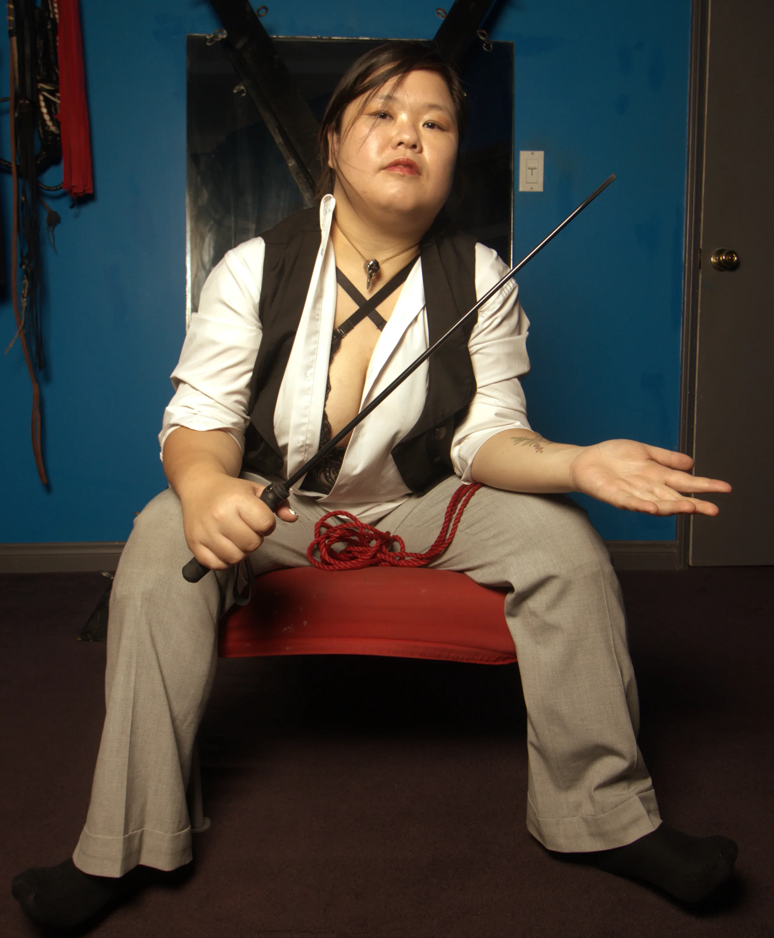 Goddess Shi (Goddess Shiney), sits in a room with BDSM equipment. She sits on a red seat, testing a black switch on her arm and contemplating.