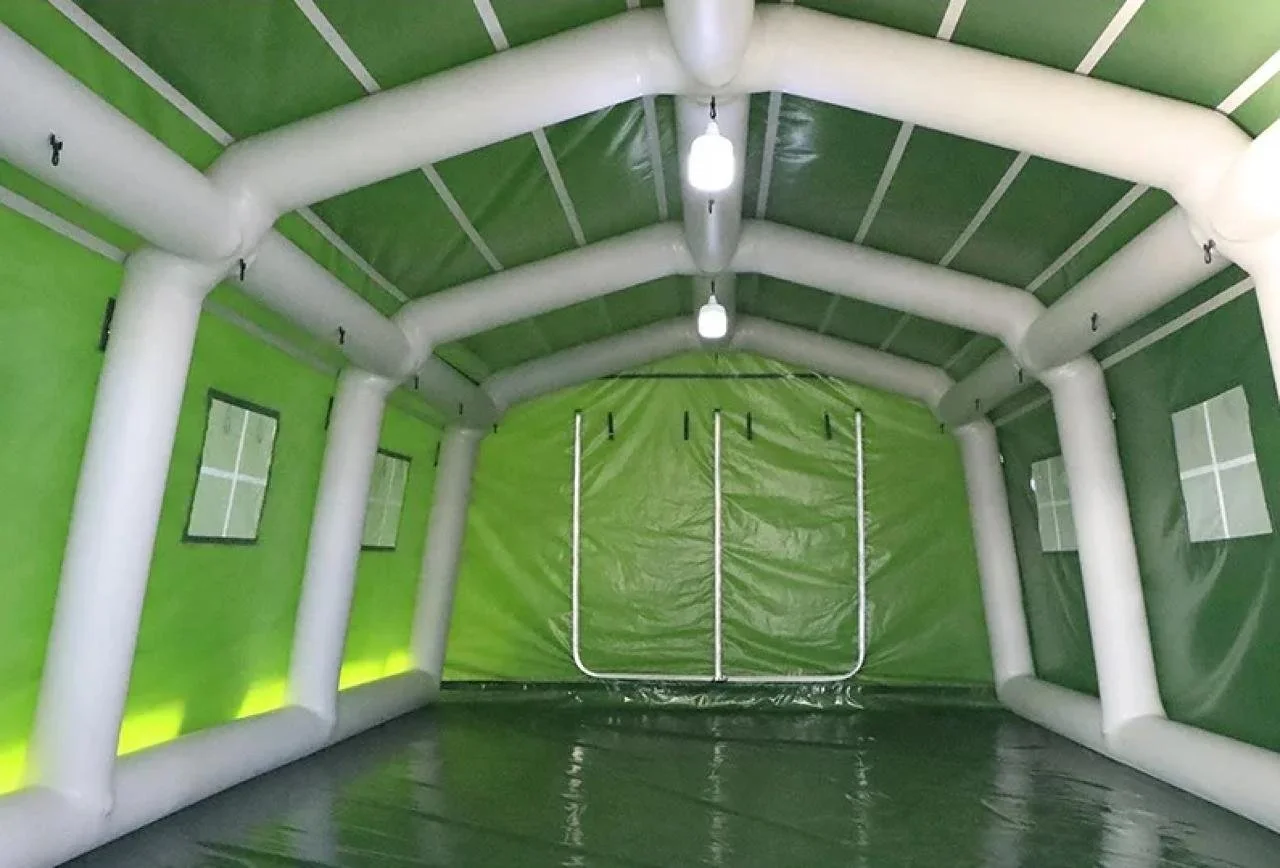 Interior of an inflated green and white party tent with window panels and a plastic floor, illuminated by hanging lights.