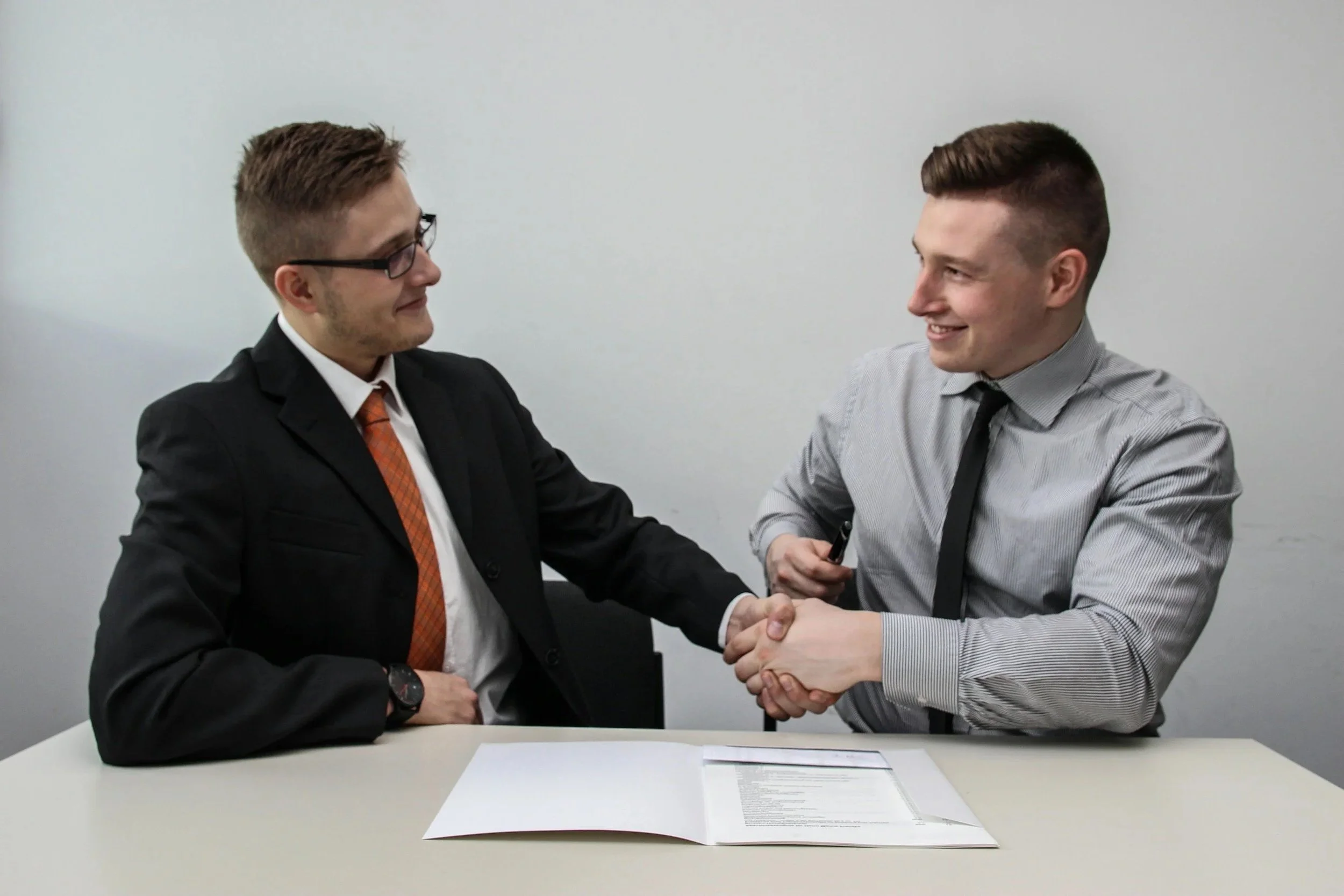Two men in business attire shaking hands and smiling in a plain white room, with an open document on the table between them.