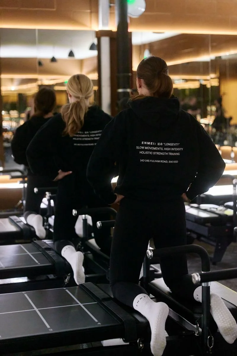 Three women are exercising on reformer Pilates machines in a gym or fitness studio. They are wearing black hoodies with text on the back, and they are performing leg lifts or similar exercises. The studio has warm lighting and mirrors, creating a professional and focused atmosphere.