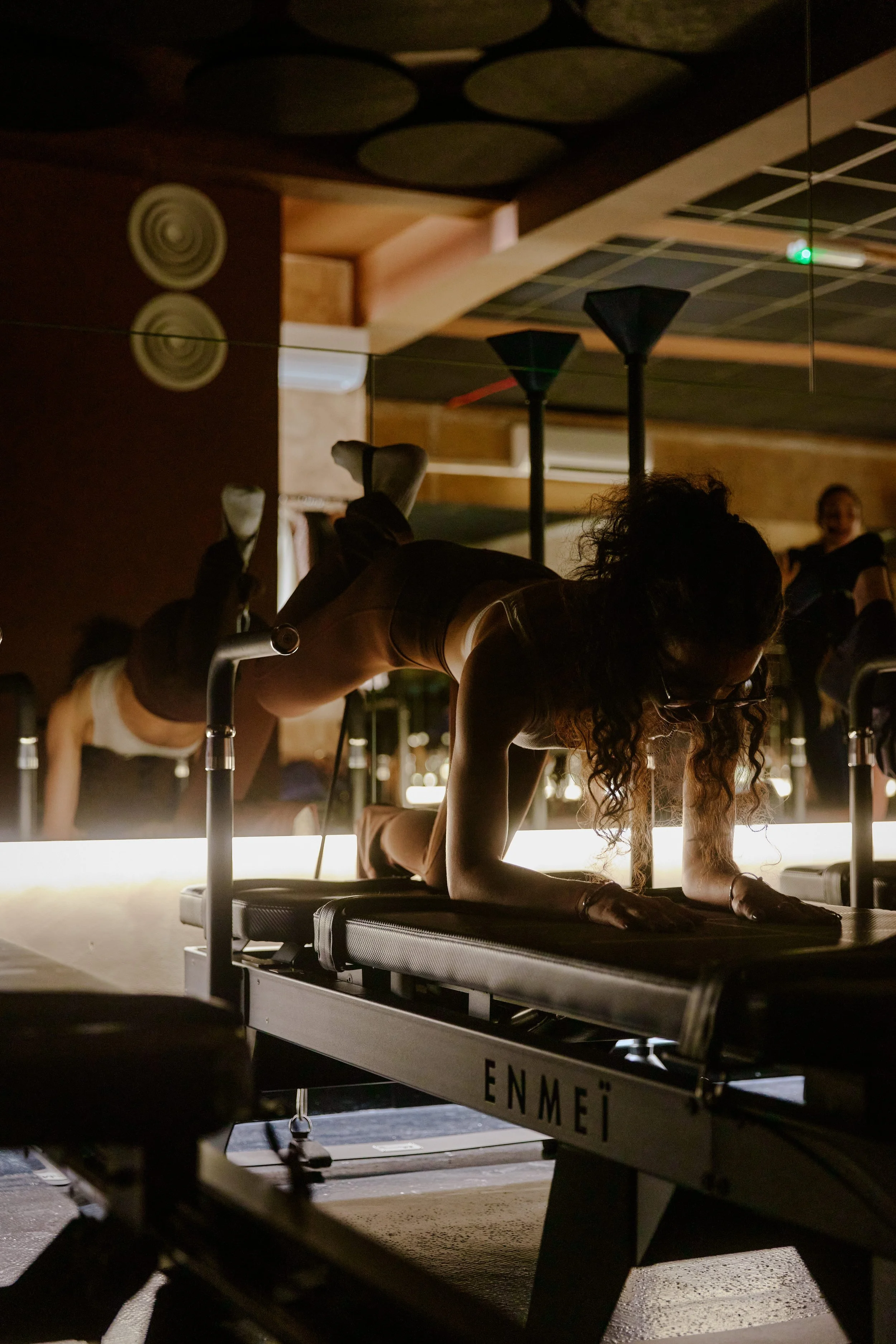 A woman with curly hair and glasses performs a Pilates exercise on a reformer machine in a dimly lit studio with warm lighting, mirrors, and other participants in the background.