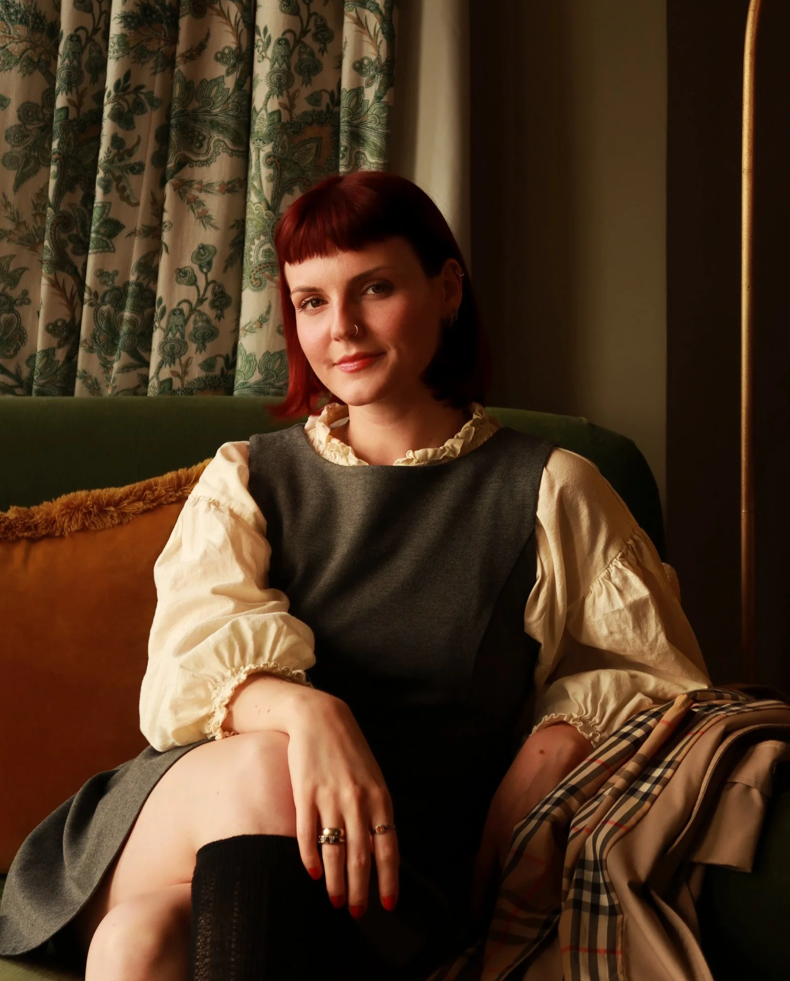 A woman with short red hair and a nose piercing sitting on a green sofa, wearing a cream blouse with puffed sleeves and a dark vest, in a warmly lit room with patterned curtains.