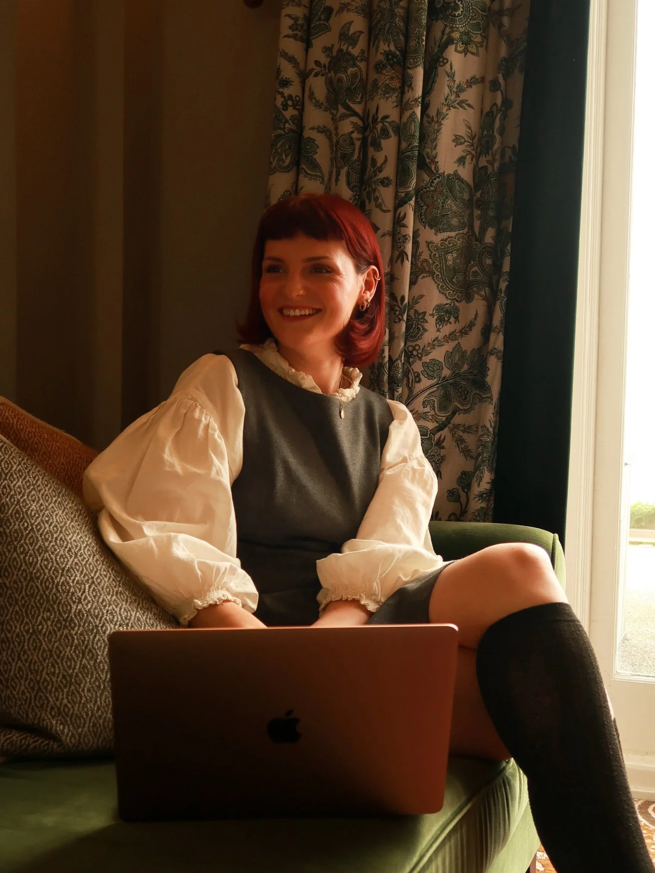 A woman with red hair and a smile sitting on a green sofa, looking to the side, with a laptop on her lap, in a room with patterned curtains and a window.