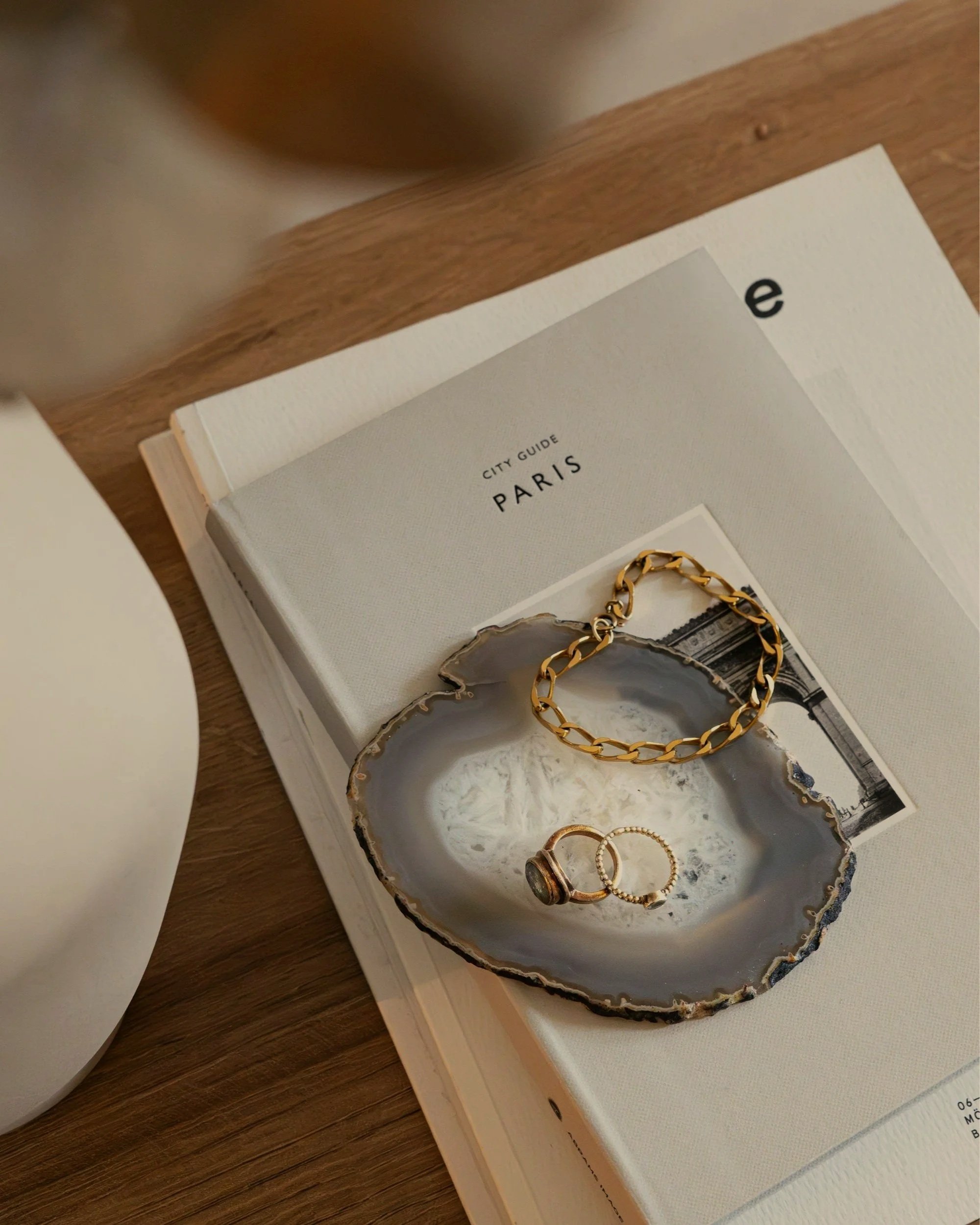 A book titled 'City Guide Paris' with a gold bracelet, pearl ring, and statement ring resting on a gray agate dish placed on the book