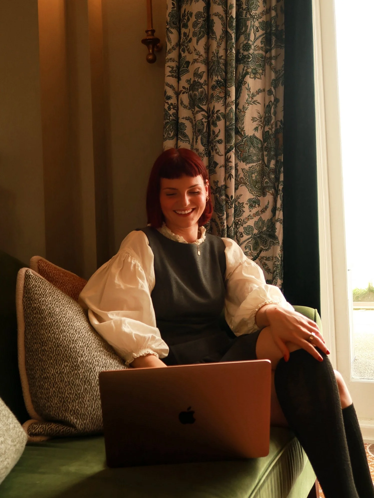 A woman with red hair sitting on a couch with a laptop on her lap, smiling, in a warmly lit room near a window with floral curtains.