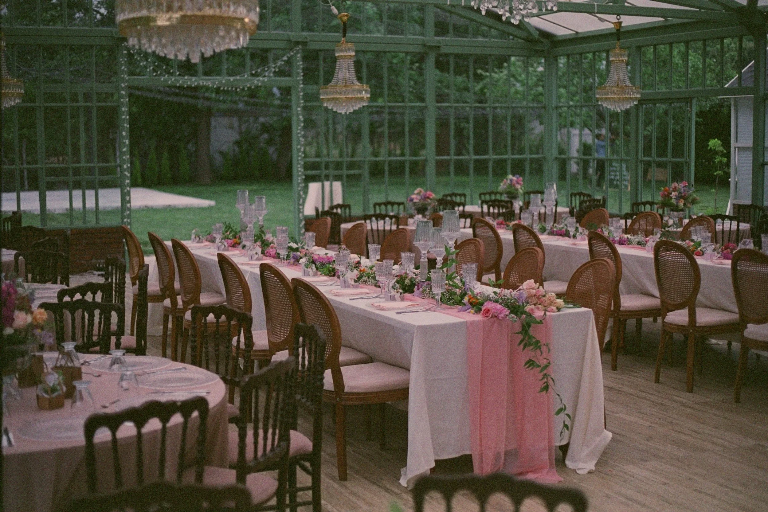 Decorated banquet tables with pink and white floral arrangements in an elegant glass greenhouse used for a wedding or event celebration.