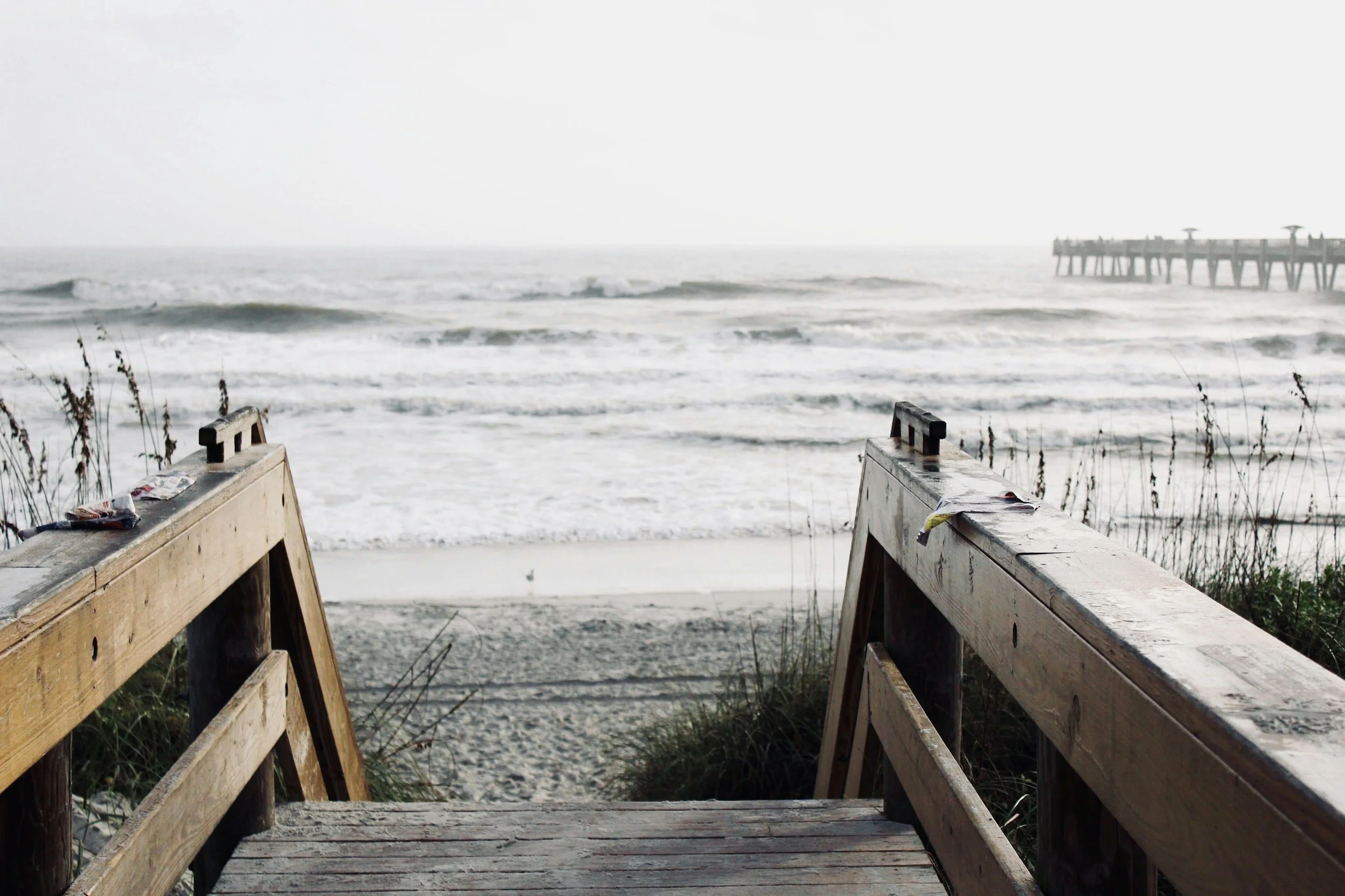View of the ocean from a wooden staircase leading down to the beach, with a pier visible in the distance on the right.