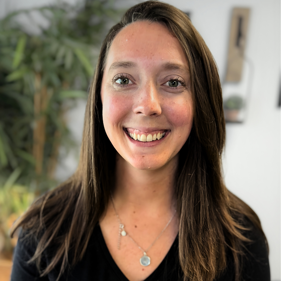 A smiling woman with long brown hair, wearing a black top and silver necklaces, against a blurred indoor background with greenery.