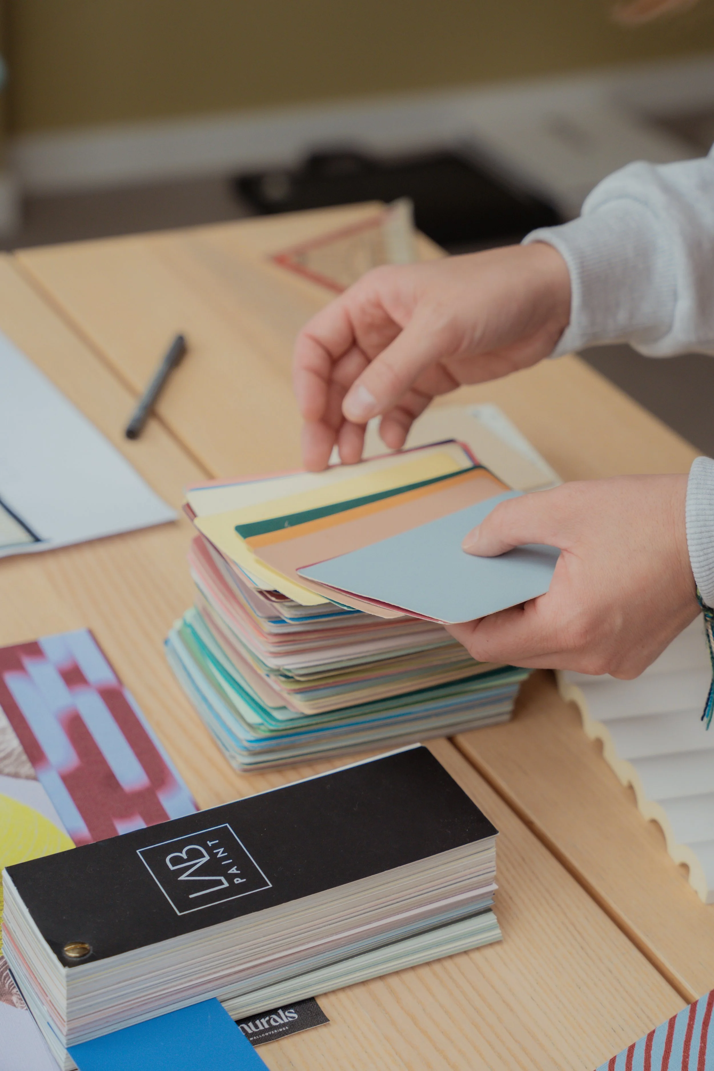 Handen die door een stapel kleurstaaltjes bladeren, op een houten tafel met ca. zes andere dozen color swatchcards, een pen en papier.