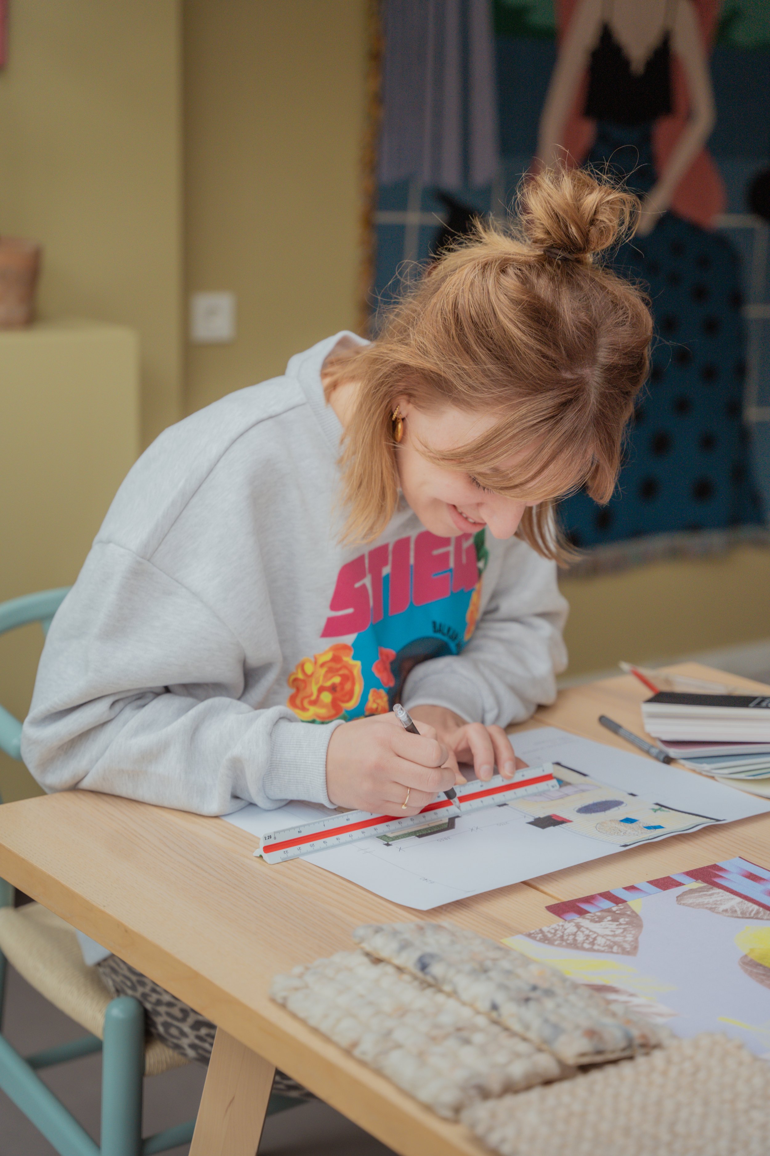 Een vrouw met bruin haar in een knot zit aan een houten tafel, schrijft met een pen op een grafiek met liniaal, op de tafel liggen boeken en kaftpapier.