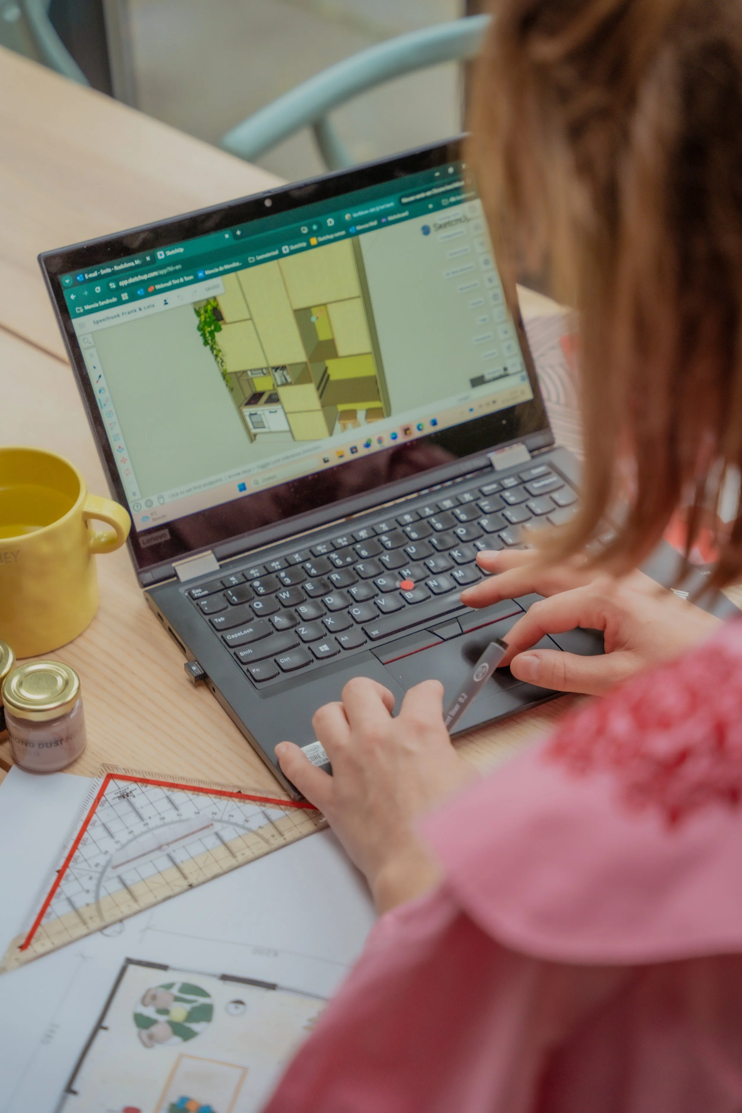 Een persoon werkt aan een laptop waarop een interieurontwerp te zien is, met gereedschap zoals een liniaal en een tekendoos op de tafel.